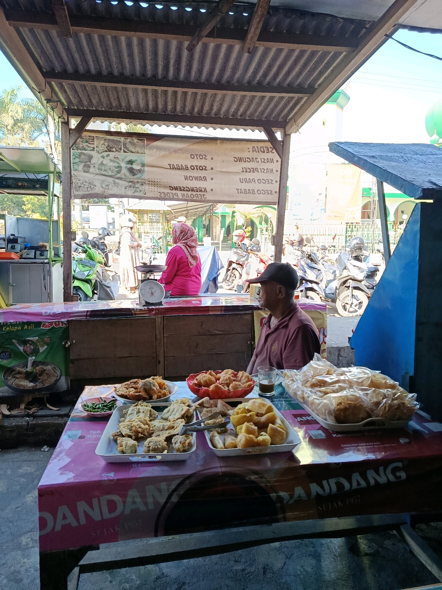 This image depicts an outdoor food stall in Bululawang, Indonesia, characterized by a corrugated metal roof supported by wooden beams, suggesting a simple, open-air structure. In the foreground, a table is laden with various food items. White platters hold an assortment of fried foods, including what appears to be tempeh and other fritters. A red bowl contains more fried items, and a small dish of green chilies is also visible. A glass of dark liquid, likely coffee or tea, sits next to a large, clear plastic bag filled with more packaged fried snacks. The table is covered with a pink and white patterned cloth that has text and logos, including "DANDANG SEJAK 1957".
A man wearing a maroon polo shirt and a dark baseball cap is seated at the right side of the table, facing left and looking away from the camera. Behind him, a woman wearing a pink hijab and a pink garment is seated on a stool next to a manual weighing scale. She appears to be tending to the stall. In the background, several motorcycles are parked, and a portion of a building with green domes and a white minaret, likely a mosque, is visible under a bright blue sky. The overall environment suggests a bustling street scene, typical of local markets or food vendors in Indonesia. The image appears to have been taken during daylight hours.