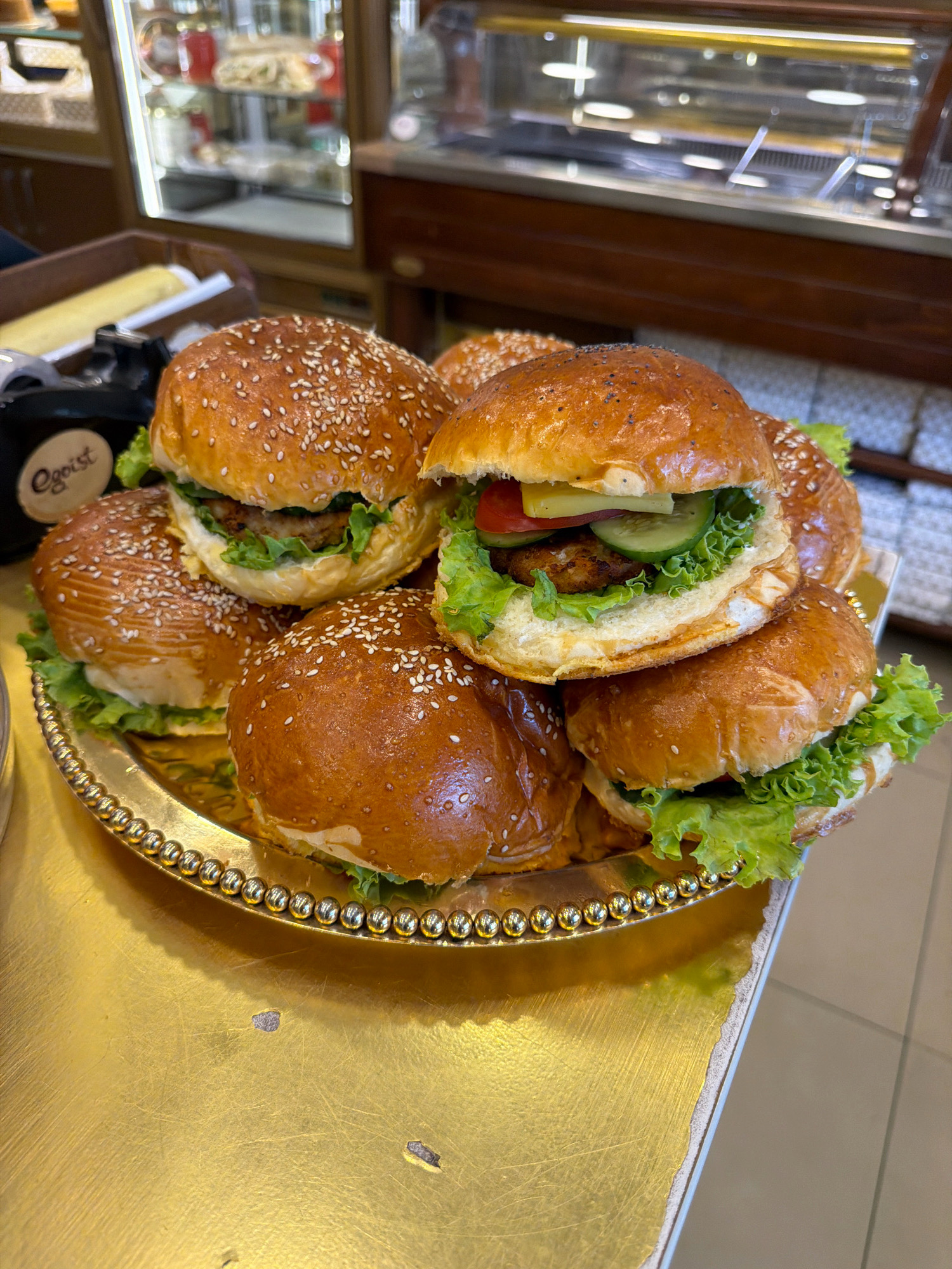 A close-up, eye-level shot shows a stack of six sesame seed-topped burgers on a golden, beaded platter. The burgers are arranged in two rows, with three on the bottom and three on top, slightly offset. The top burger is the most visible, revealing its contents of lettuce, a patty, tomato, cucumber, and cheese. The platter sits on a golden surface, likely a table, with a blurred background of what appears to be a bakery or cafe display case filled with food items. The overall scene is brightly lit, with the golden tones of the platter and table dominating the foreground. This image was taken in Baku, Azerbaijan.
