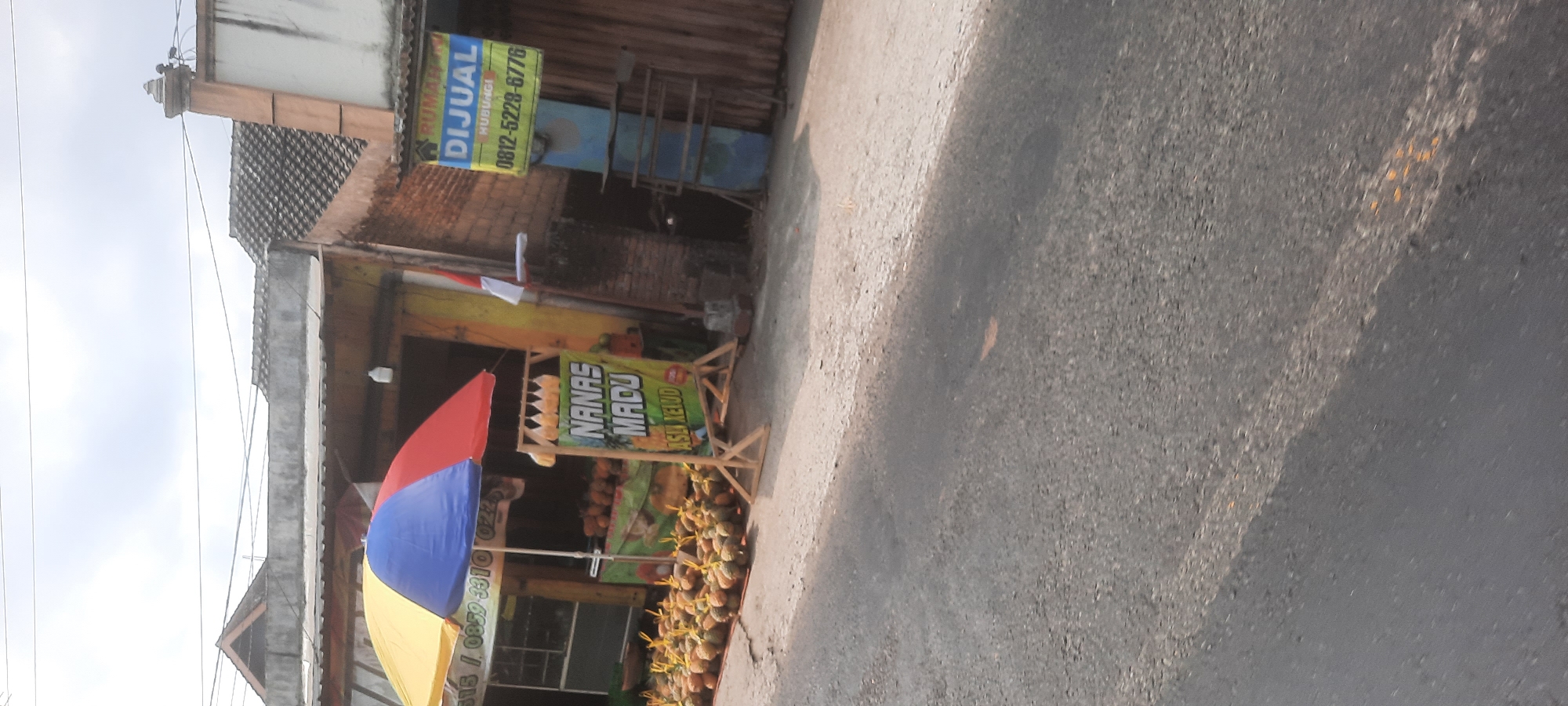 The image depicts a roadside stall in the city of Kesamben, Indonesia, where pineapples are being sold. In the foreground, a large pile of pineapples is arranged on the ground, some of which are tied with string. Behind this, a stall displays more pineapples, with a sign that reads "NANAS MADU ASLI KELUD". A colorful umbrella, with segments of yellow, blue, and red, provides shade over the stall. To the right of the stall, a blue metal structure and a corrugated metal wall are visible. Above this, a banner with the word "DIJUAL" (for sale) and a phone number "0812-5228-6776" is prominently displayed, indicating a property sale. The background shows buildings with tiled roofs, typical of the Indonesian architecture. The overall scene suggests a commercial area on the side of a road, with a focus on agricultural produce.