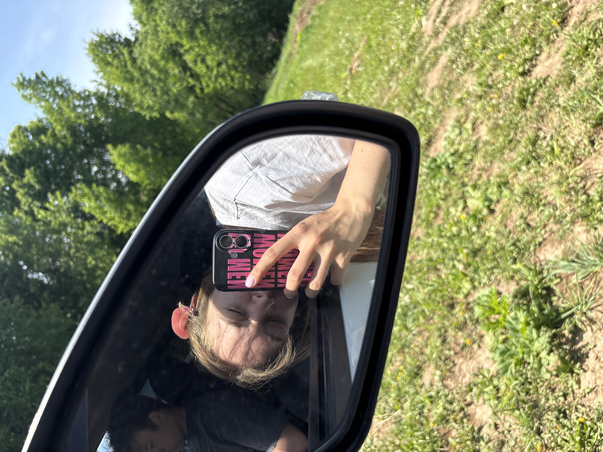 A side-view mirror reflects a young individual holding a phone to take a picture of themself. The phone case is black with pink letters. Their eyes are closed. The reflection also reveals a partial view of the person seated inside the vehicle. Outside the car, blurred vegetation is visible. The location is not discernible.