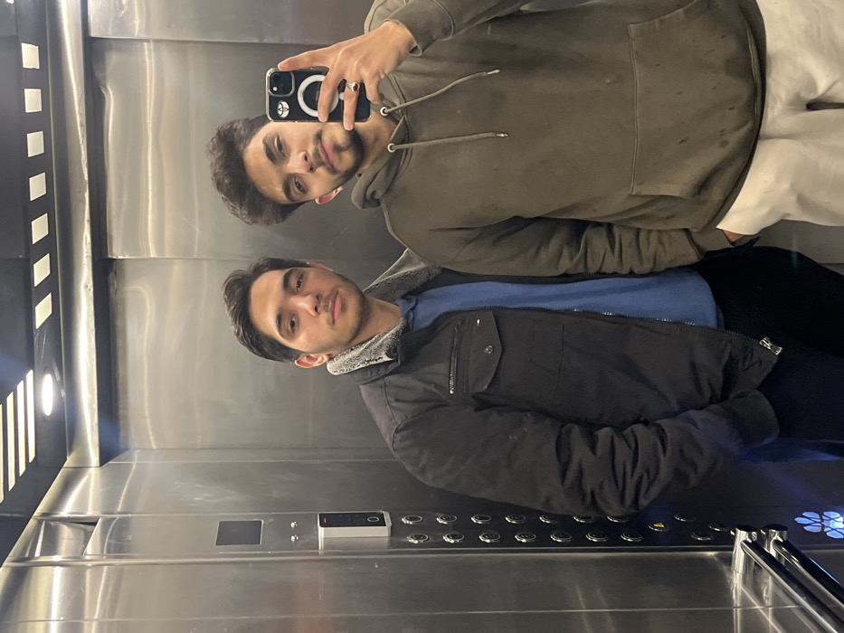 Two young adult males are depicted inside a brushed stainless steel elevator, located within Kafr Sousa Municipality, Syria. The elevator interior features reflective walls, a control panel with numerous circular buttons and a vertical digital display on the left side, and recessed rectangular and linear light fixtures on the ceiling. The male on the viewer's left has short dark hair and a light beard, wearing a dark jacket with a light-colored, possibly faux-fur, collar lining over a blue long-sleeved shirt. His left hand is partially obscured, possibly in a pocket. The male on the viewer's right has curly dark hair and a light beard, wearing an olive-green hooded sweatshirt with a front pouch pocket and light-colored trousers. He is holding a dark-colored smartphone, equipped with a white circular accessory on its back, in his right hand, angled to capture their reflection in a mirror selfie. A dark ring is visible on his left ring finger. Both individuals are looking directly towards the camera's perspective.