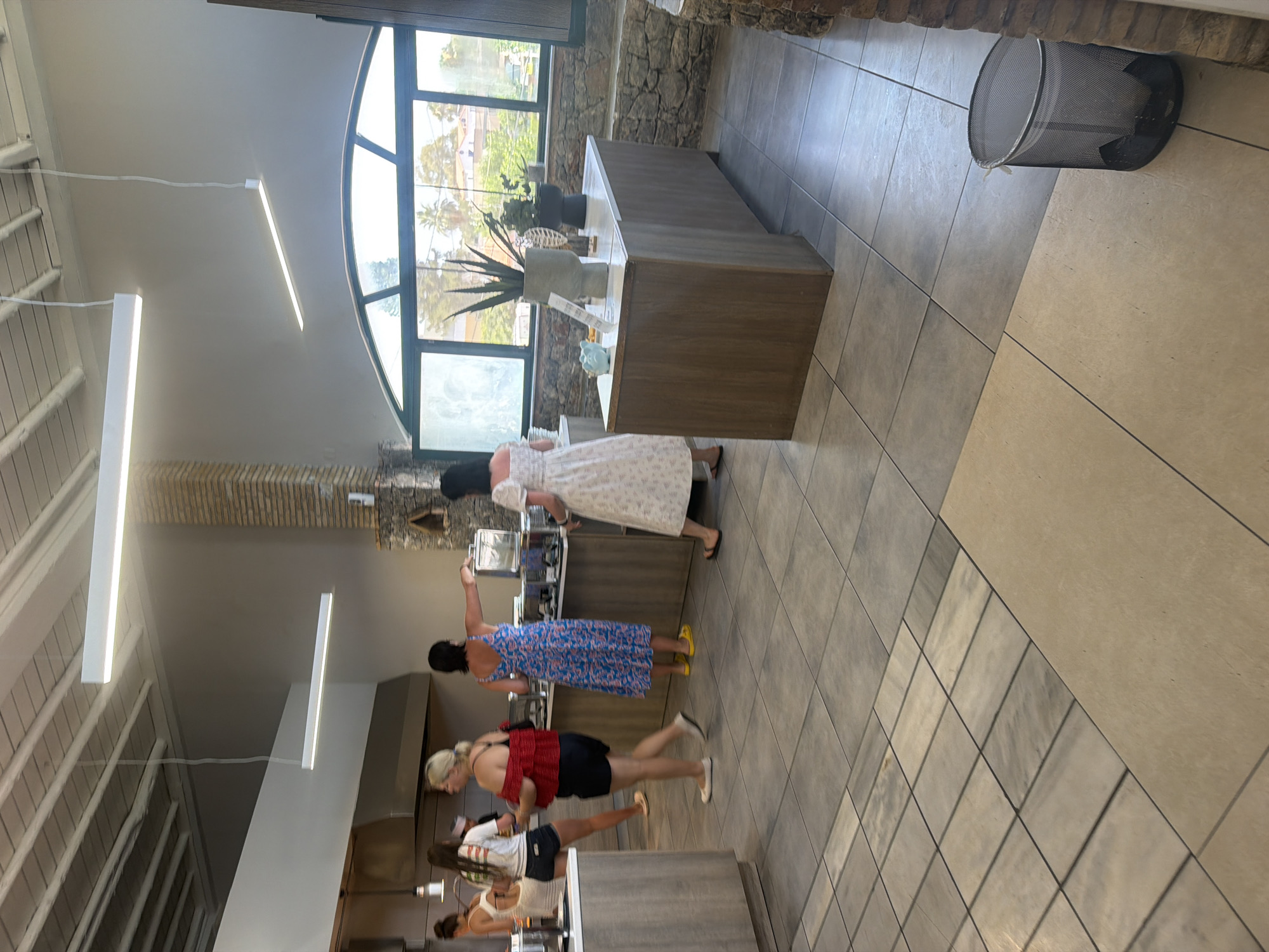 This image depicts the interior of a restaurant in Georgioupoli, Greece, featuring a buffet-style food service area. Several women are visible, some interacting with the buffet and others walking through the space. The architecture includes a tiled floor, a brick pillar, and a large arched window offering a view of outdoor greenery. Modern overhead lighting fixtures illuminate the dining area.