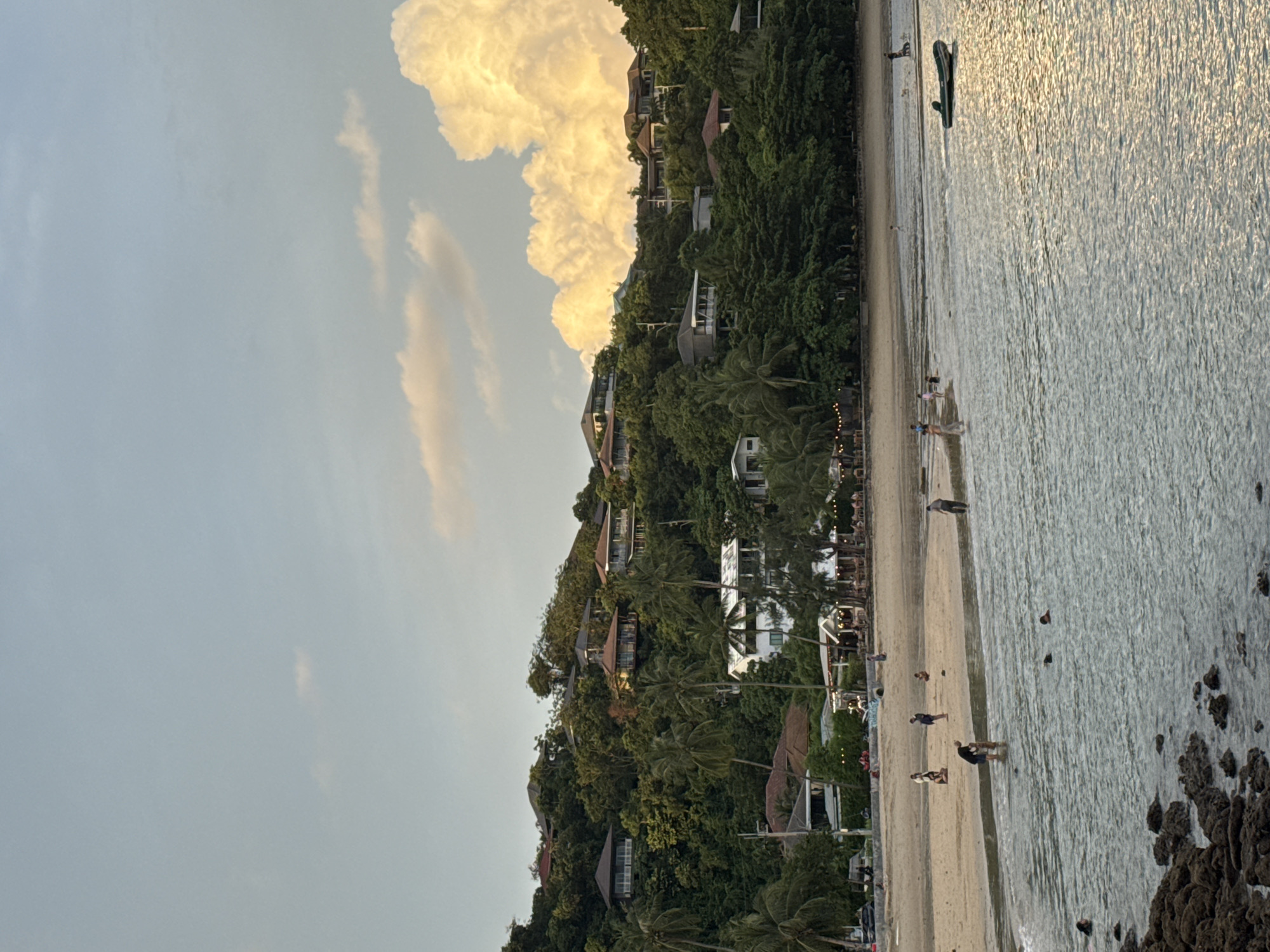 This image depicts a tranquil beach scene at sunset in Wichit, Thailand. A gentle slope covered in lush green trees rises from the sandy shore, dotted with several houses nestled among the foliage. The sky above is a soft blue, with a few wispy clouds and one large, illuminated cumulus cloud bathed in the warm glow of the setting sun.
On the beach, a few people are scattered about. Some are walking near the water's edge, while others are closer to the buildings. In the foreground, the calm sea reflects the sky and the golden light. A small inflatable boat is moored near the shore, its dark green hull visible against the shimmering water. Rocky outcrops are present in the lower left corner of the image. The overall atmosphere is peaceful and serene, capturing the relaxed ambiance of a tropical evening.