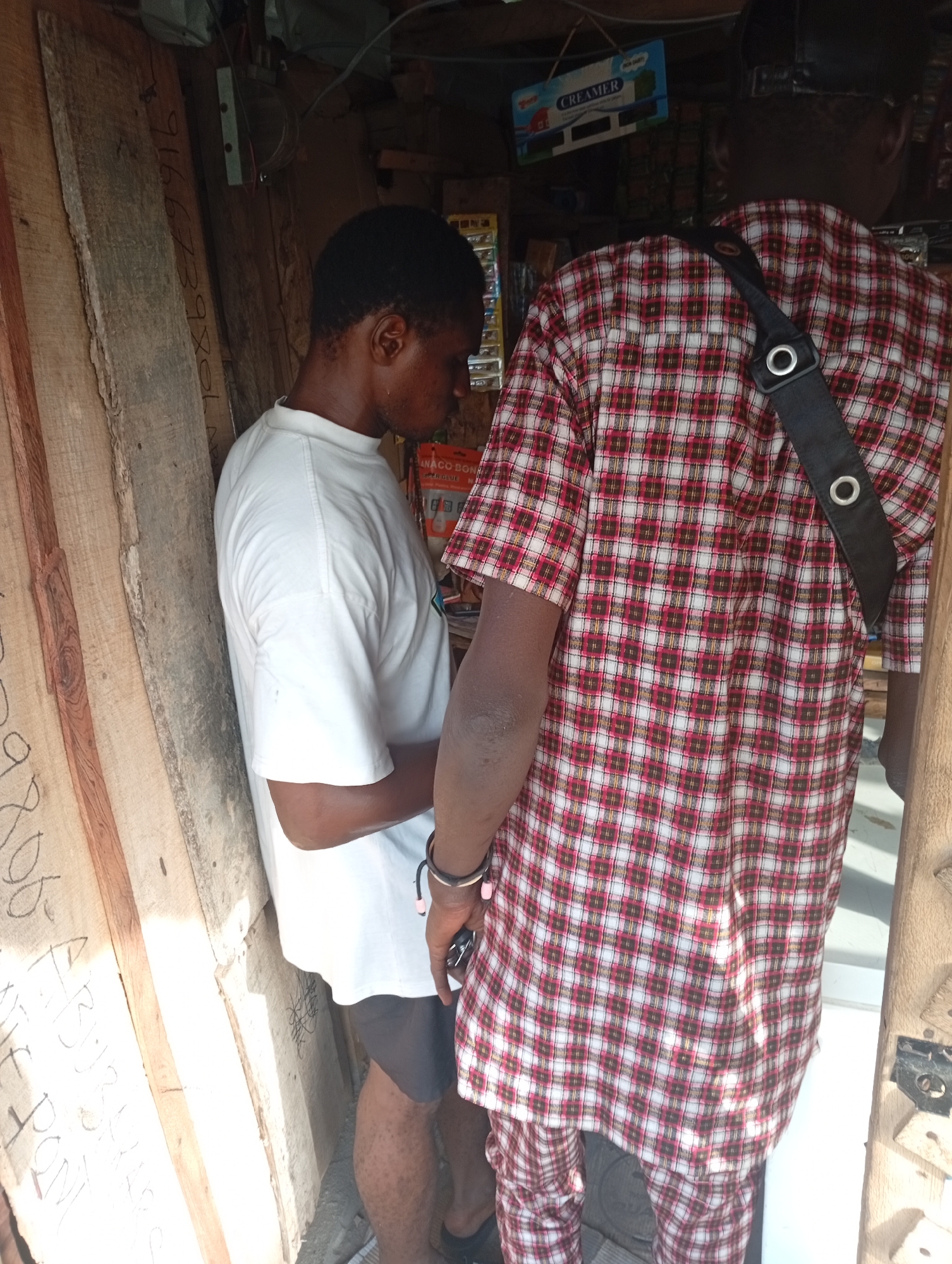 Two men are standing inside a shop in Adankolo, Nigeria. One man is wearing a white t-shirt and dark shorts, and is looking down, holding a dark object in his hand. The other man is wearing a red and white plaid shirt and matching pants, with a black shoulder bag. The shop has wooden walls and shelves filled with various items, including a "CREAMER" brand product.
