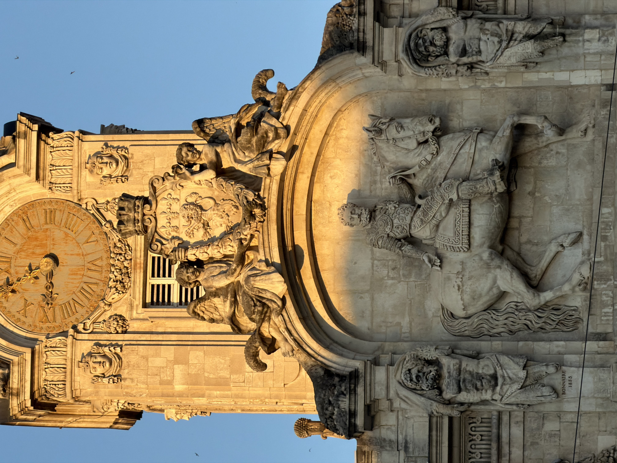 The image captures an upward view of a stone facade adorned with elaborate sculptures, likely a historical building in Lyon, France. The central element is a large equestrian statue of a man in ornate armor, possibly a historical figure, depicted in profile with his horse. Flanking this central piece are two imposing, muscular male figures carved in stone, wearing draped garments and animal pelts over their heads. Below the equestrian statue, the name "BONNAIRE" and the year "1855" are inscribed, indicating the sculptor and date of creation. Above the equestrian statue, two winged figures, possibly cherubs or angels, are seated. The entire composition is set against a clear blue sky. The lower portion of the image shows the finely detailed stonework of the building's facade, including decorative carvings and a window with louvers. The golden light suggests it is either sunrise or sunset, casting long shadows and highlighting the textures of the stone.