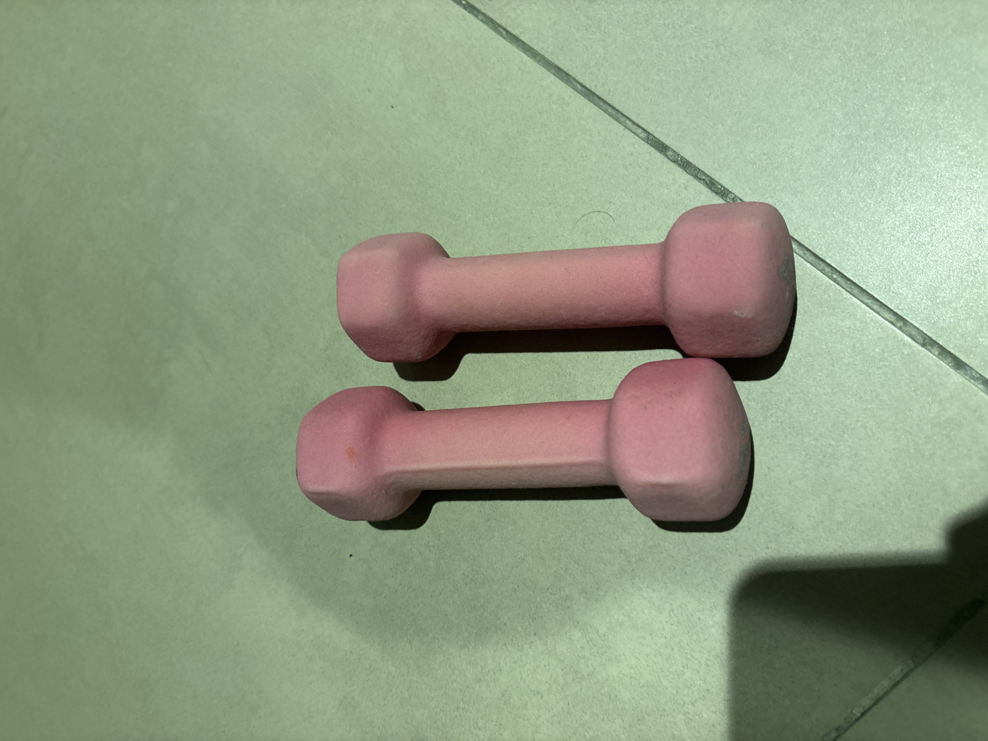 Two pink dumbbells are lying on a tiled surface. The dumbbells are identical in shape and size, with hexagonal ends and a cylindrical grip. They are positioned side-by-side, with the left dumbbell slightly closer to the foreground than the right. The tiled surface appears to be light green and has a grid pattern formed by grout lines. Shadows are cast by the dumbbells, suggesting overhead lighting. No people or other objects are visible in the image. The scene is devoid of any context that would place it in Dubai, United Arab Emirates.