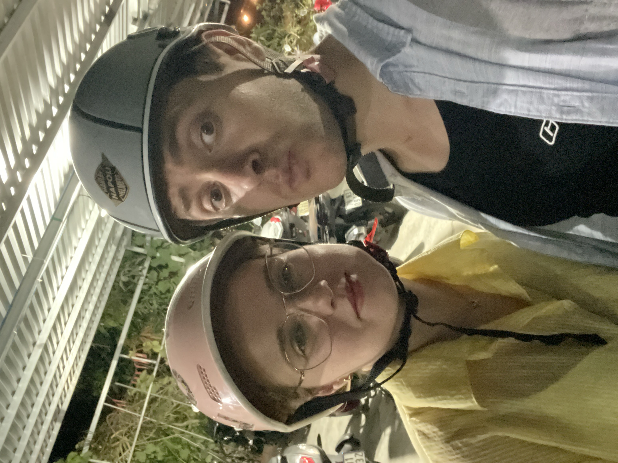 A selfie-style photograph, rotated counter-clockwise, shows two individuals wearing helmets. The person on top wears a gray helmet with a white stripe and a "Napoli" emblem. They have light skin, dark hair, and are making a pouting facial expression. The person below wears a pink helmet, glasses, and a yellow shirt. Both are secured with black helmet straps. The background shows glimpses of other motorcycles, vegetation, and a white metal fence. Based on the metadata, this photo was taken in Diên Khánh, Vietnam.