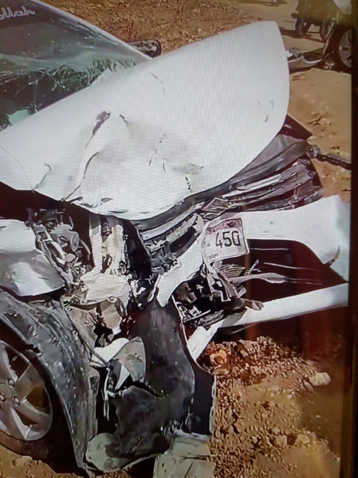 The image depicts the aftermath of a severe car crash. A white car is heavily damaged, with crumpled metal, broken windshield, and a missing front end. The license plate is visible, showing "450" with "REM" underneath. The car rests on dirt, indicating an off-road location, possibly in Unknown, Unknown. The image suggests a high-impact collision, likely involving a single vehicle.
