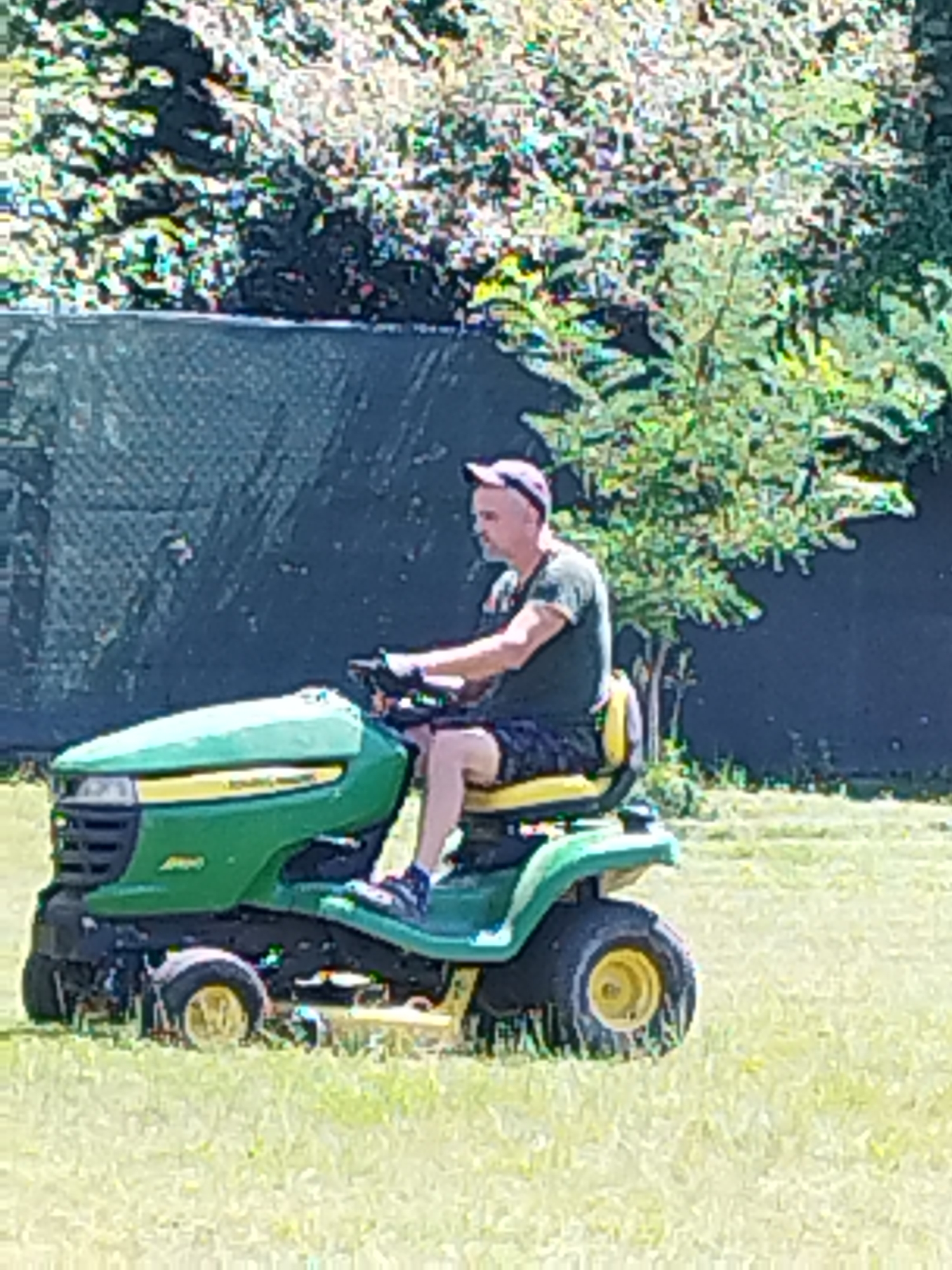 A man with a light complexion and a shaved head is operating a green John Deere riding lawn mower. He is wearing a dark green t-shirt, dark shorts with a patterned design, black gloves, and a pink baseball cap. The man is positioned on the seat of the lawn mower, holding the steering wheel with both hands and looking forward. His left foot is on the mower's footrest, and his right foot is on the ground. The lawn mower is on a grassy lawn, with a dark green fence and lush green trees in the background. The lighting suggests it is daytime, with sunlight illuminating the scene. The image does not contain any specific indicators of its location within Bucharest, Romania.