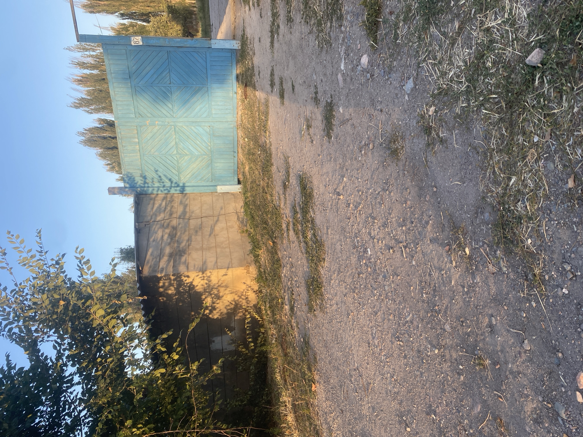 The image shows a dirt path leading to a light blue double gate. To the left of the gate is a concrete wall, partially shaded by trees. The gate has a geometric pattern on it and a small white sign with the number "67" on it. Tall, thin trees line the background. The ground is covered with dirt and sparse dry grass. The scene appears to be in a rural or semi-rural area.