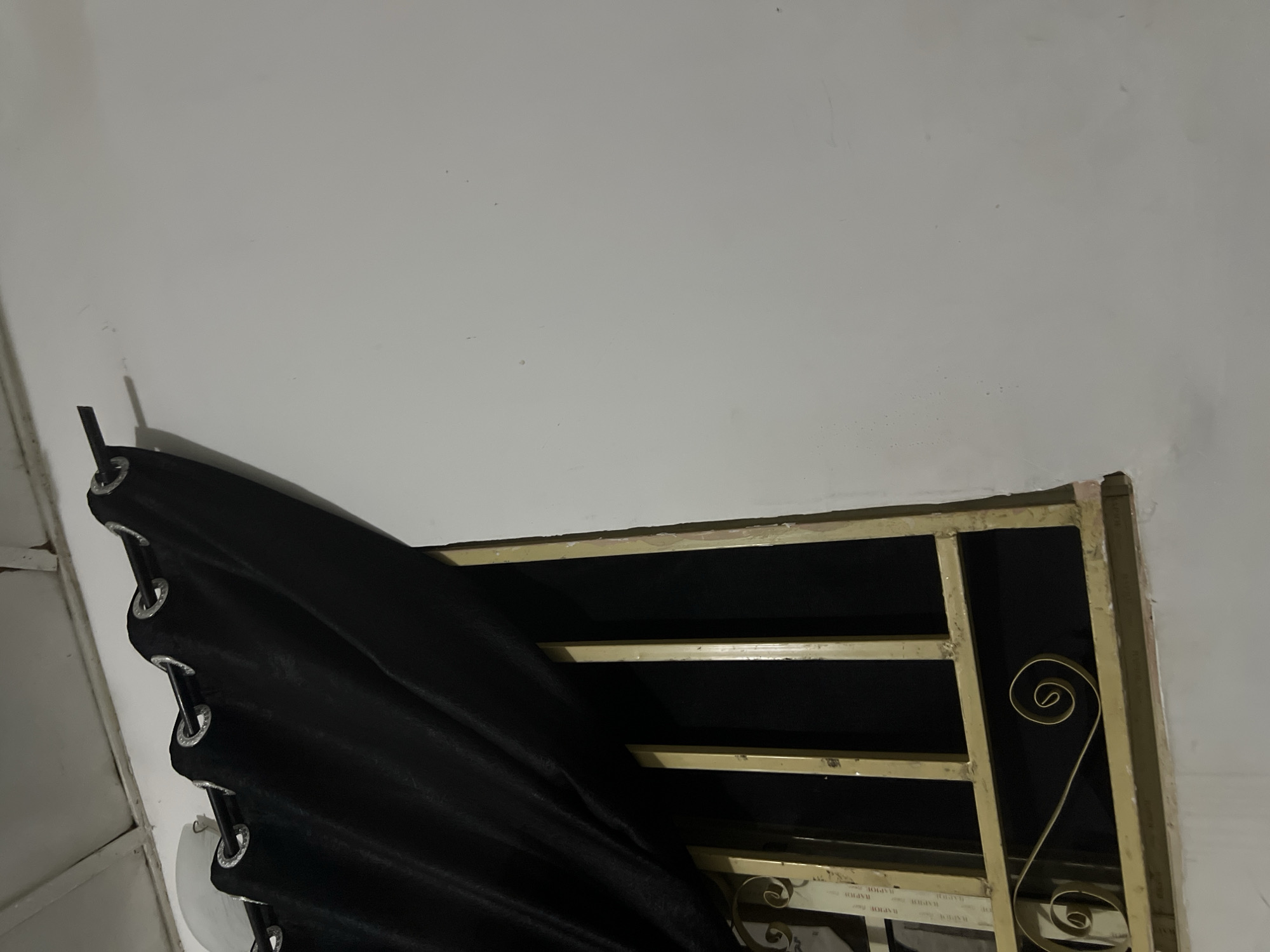 The image shows a close-up view of a window with black curtains partially drawn across it. The window is covered by a metal grate painted gold. The wall next to the window is painted white. The ceiling, visible at the top of the image, is also white and appears to be made of paneling. The lighting in the scene is dim, suggesting it may be nighttime or indoors with artificial light. The image is taken from a low angle. The location is identified as Howayi, Nigeria.