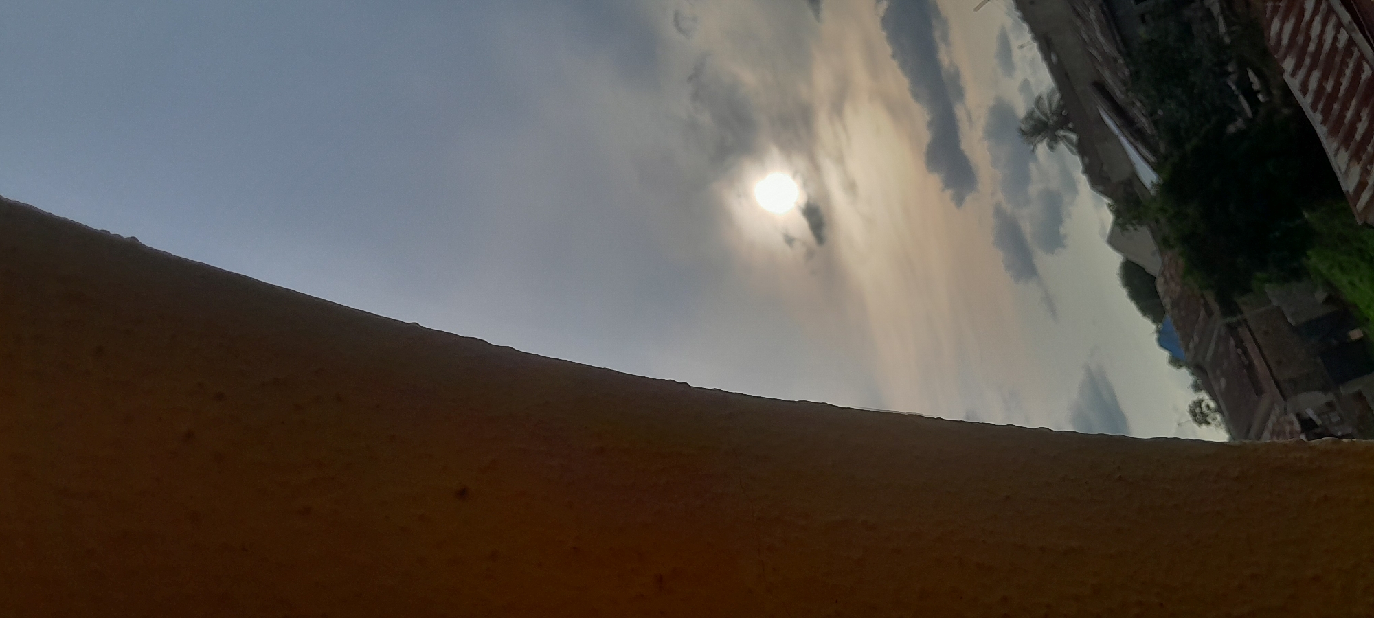 This is a vertical, eye-level, medium shot of a cloudy sky with the sun partially obscured. The sun, a bright white circle, is positioned in the upper right quadrant of the image. Dark gray clouds are scattered across the sky, with lighter gray clouds present near the sun. In the lower right portion of the frame, the roofs of several buildings are visible, appearing to be a mix of corrugated metal and some lighter colored materials. A palm tree is also visible behind the buildings, its fronds silhouetted against the sky. The lower left portion of the image is dominated by a textured, curved surface, which appears to be a wall or architectural feature, casting a shadow that occupies roughly the left third of the frame. The overall lighting suggests either early morning or late afternoon. This scene is consistent with a residential area in Abuja, Nigeria.