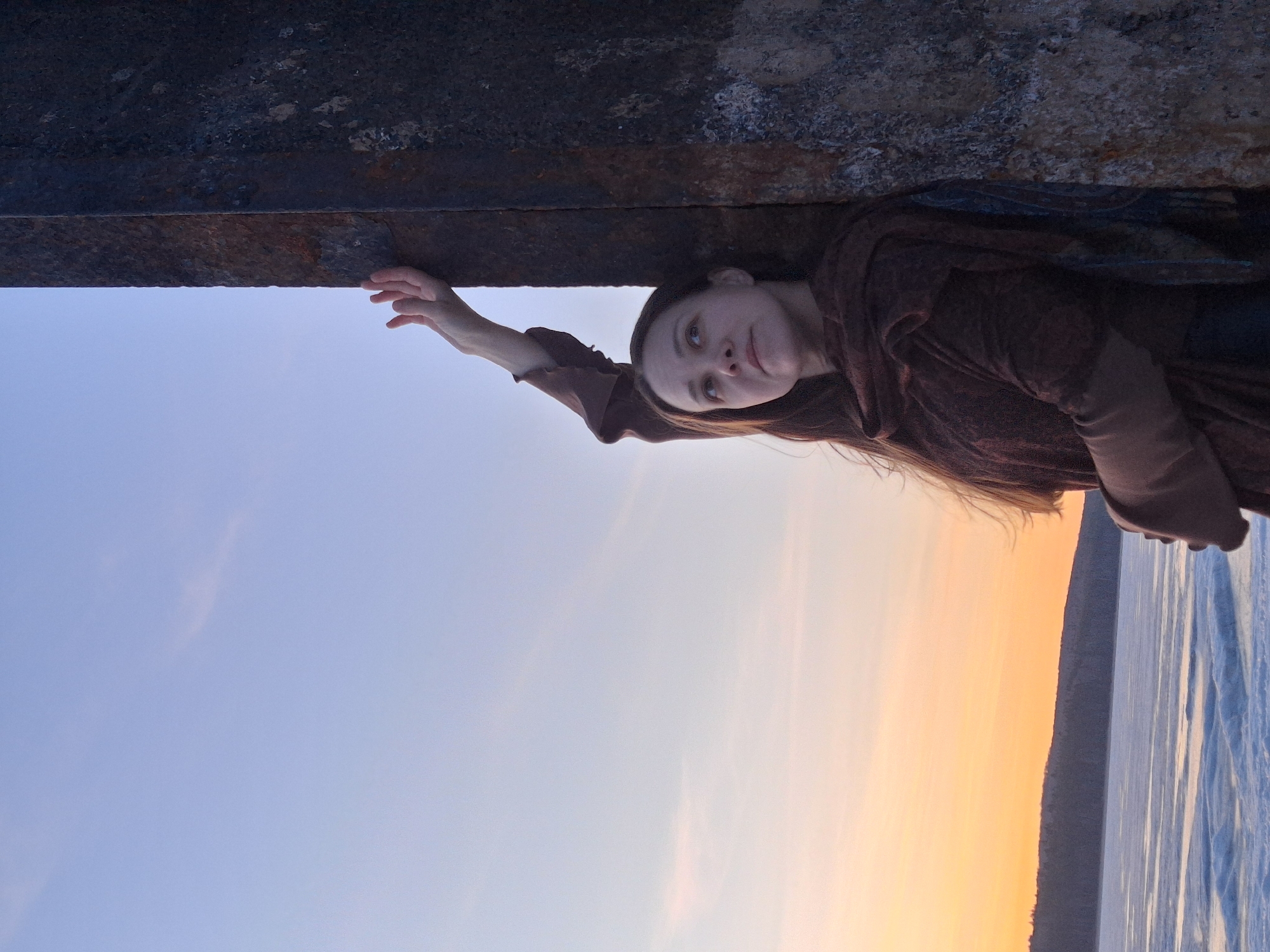 A young woman with long, dark hair is depicted in a serene outdoor setting. She wears a dark, possibly brown or maroon, long-sleeved garment. Her right arm is extended upwards, with her fingers lightly touching the underside of a large, weathered horizontal beam, which appears to be made of rusty metal or concrete. She looks upwards and slightly to her left (viewer's right), exhibiting a calm and introspective expression.
The scene is set against a vast sky that transitions from a soft blue on the left to warm orange and pink hues on the right, strongly suggesting either sunrise or sunset. Below the woman and the beam, a wide expanse of what appears to be water or a frozen body of water (ice) stretches into the distance, reflecting the sky's colors. A dark, low-lying landmass or distant hills are visible on the horizon, separating the water from the sky. The overall atmosphere is tranquil and contemplative. There is no visible text in the image. The location context provided indicates this scene is in Chaikovsky, Russia.
