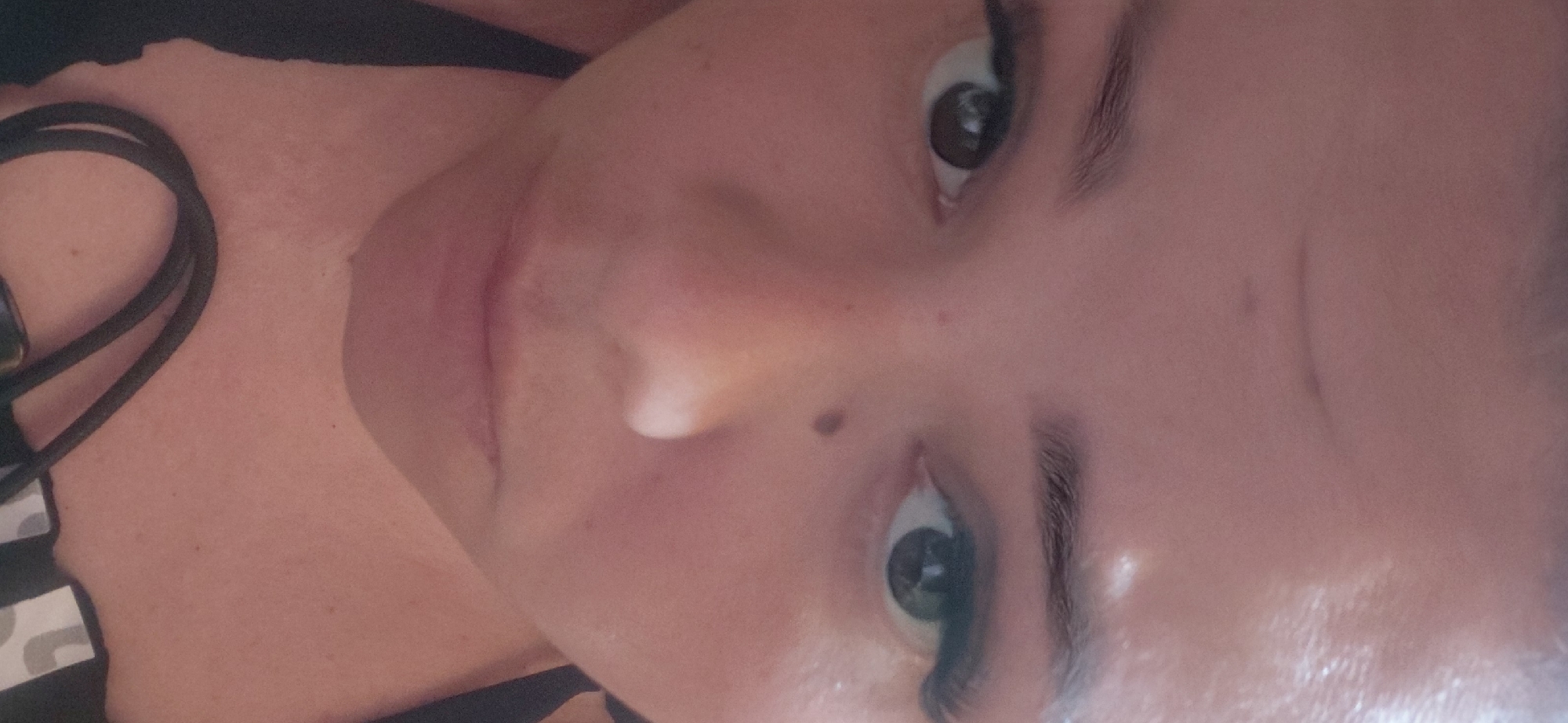The media file is a close-up, vertically oriented photograph of a person's face and upper chest. The subject appears to be a young woman, with fair skin, dark eyebrows, and large, dark eyes. She is wearing what looks like dark clothing around her neck and shoulder. One of her eyes is clearly visible, looking directly at the camera. Her lips are slightly parted, and her skin has a smooth texture, with a mole visible on her cheekbone.
The lighting suggests an indoor setting, possibly with artificial light, as there are some shiny highlights on her skin. The overall mood is intimate due to the close-up framing. The background is largely out of focus, but the dark material around her neck, possibly a strap or collar, and the hint of patterned fabric on her shoulder are discernible. The image is candid and personal, focusing on the subject's features. There is no visible text in the image. The photograph is cropped tightly on the subject's face, with a portion of her neck and upper chest visible.