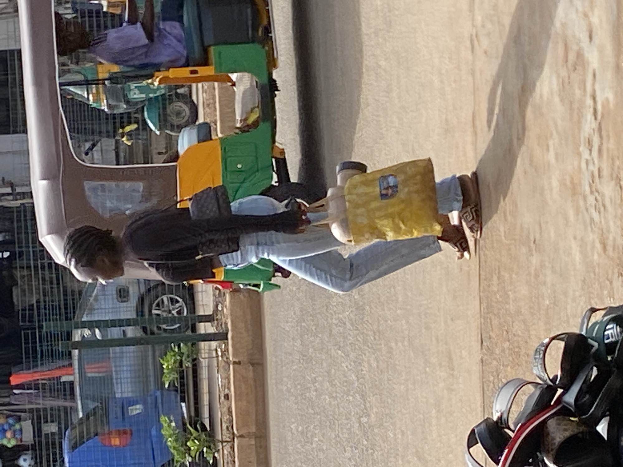 The image depicts an outdoor street scene in Bukuru, Nigeria, illuminated by bright daylight, casting strong shadows. The central subject is a person, likely a young adult, standing on a paved road or sidewalk. They are dressed in light blue jeans, a dark long-sleeved top, and brown sandals, carrying a yellow shoulder bag and another dark bag. Their hair appears to be styled in braids or dreadlocks.
In the mid-ground, several auto-rickshaws, known locally as kekes, are visible. One prominent keke features green and yellow panels, with a passenger seated inside wearing a light purple shirt. Another keke with blue and lighter-colored panels is partially seen. The background shows a fence, behind which parts of a white or light-colored car are visible, along with some green foliage and a blue tarp or fabric. In the immediate foreground, to the bottom right, is a display of various sandals, suggesting a roadside vendor or market stall. The strong sunlight and distinct shadows indicate it is daytime, likely morning or late afternoon. No discernible text is clearly visible on any objects.