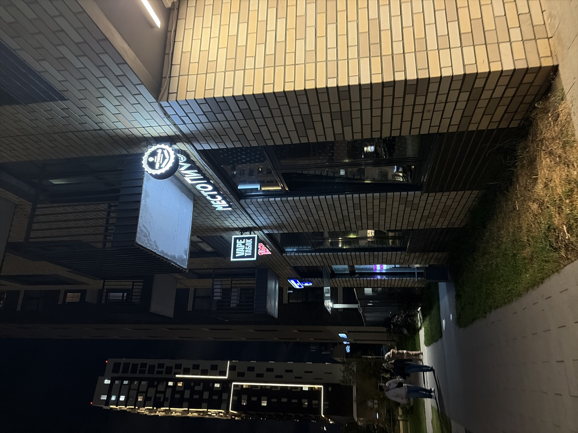 The image depicts a nighttime street scene in Moscow, Russia, featuring a modern brick building with illuminated signage. In the foreground, a paved sidewalk runs alongside a grassy area. A tall, multi-story building with a distinctive vertical light design is visible in the background. On the side of the brick building, several neon signs are displayed. One circular sign with a white illuminated emblem reads "МЕСТО ПИВ" (Mesto Piv), suggesting a place for beer. Below it, a rectangular sign in Russian reads "VAPE ТАБАК" (Vape Tabak), indicating a vape and tobacco shop. Two individuals, a man and a woman, are walking on the sidewalk, appearing to be engaged in conversation. The overall atmosphere is urban and contemporary, illuminated by artificial lights typical of a city at night.