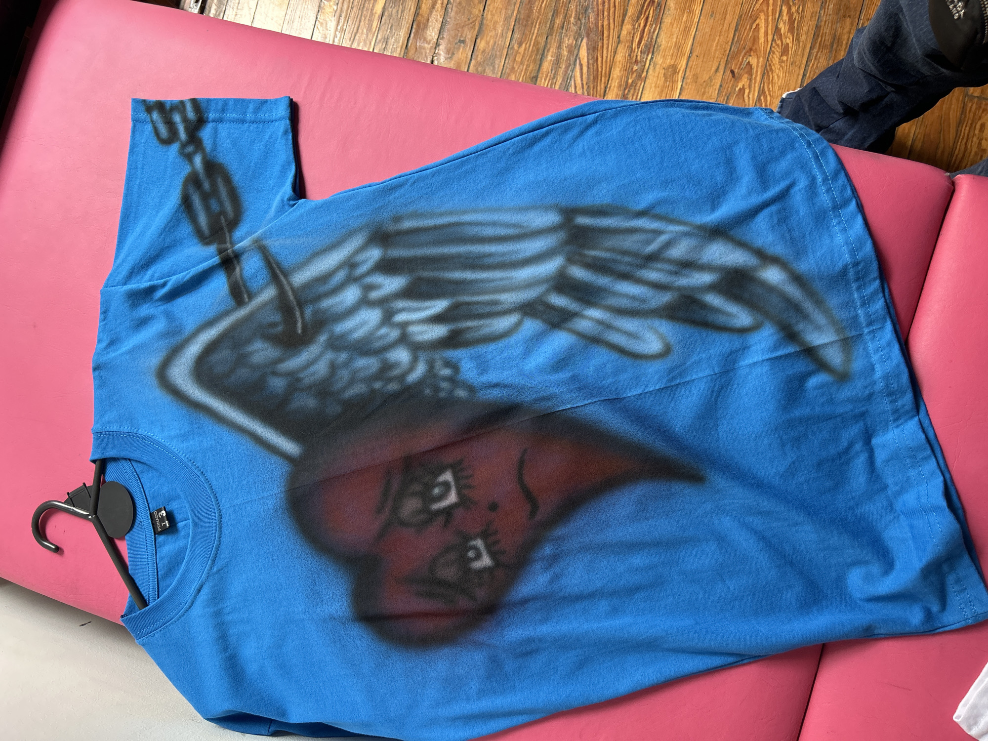 A blue t-shirt with an airbrushed design of a heart with wings and a chain hangs on a black plastic hanger on a pink padded surface. In the background, a section of wooden floorboards is visible, along with a dark blue fabric. The location of the image cannot be determined from the available information.