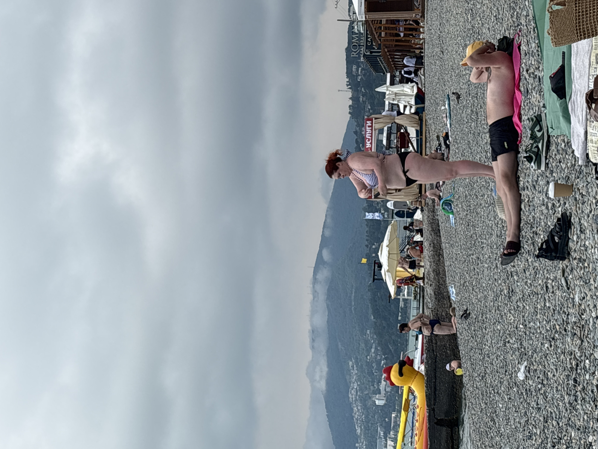 The image depicts a cloudy day at a pebble beach in Sochi, Russia. In the foreground, a man wearing a yellow cap and black shorts lies on a pink towel, his hands behind his head. To his left, a woman in a striped bikini stands, adjusting her clothing. In the background, several beach umbrellas and structures, including one with "ROM" visible, suggest a resort area. A large yellow inflatable duck is visible on the left side of the image, near the water's edge. Mountains covered in mist and trees form the backdrop, with buildings nestled on their slopes. Other beachgoers are scattered across the scene, some in the water and others relaxing on the shore. The overall atmosphere is relaxed and casual, typical of a beach day, despite the overcast sky.