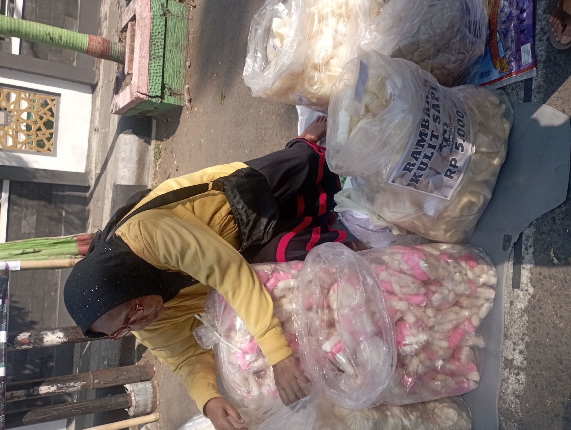 A person wearing a yellow long-sleeved shirt, a black hijab, and glasses is tending to several large plastic bags filled with snacks. The person is positioned on the right side of the frame, leaning forward and reaching into one of the bags. The snacks appear to be light-colored, with some having pink accents. One bag has a label that reads "RAMBA KULIT SAPI RP 5.000". The scene is outdoors, with a paved surface and a concrete curb visible. In the background, there is a building with a patterned window and a tree in a planter. The lighting suggests it is daytime, with shadows cast on the ground. The overall impression is a street vendor selling snacks in Nganjuk, Indonesia.