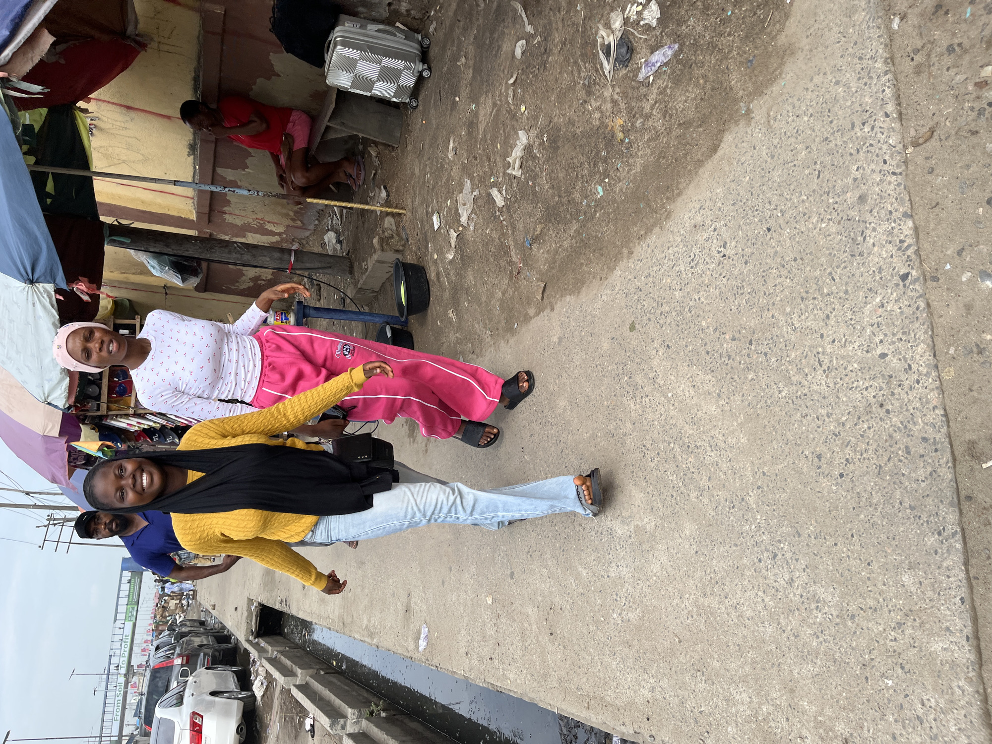 Two women are posing for a photo on a sidewalk in Lekki, Nigeria. The woman closest to the camera is wearing a yellow sweater, light blue jeans, a black head covering, and sandals. The other woman is wearing a white top, pink pants with white stripes, a pink head covering, and sandals. They are both smiling. Behind them is a busy street with cars, and a man in a blue shirt is partially visible. In the background, there is a building with a red awning and another person sitting on the ground. There is a suitcase near the building. Trash and debris are scattered on the ground near the building. The sidewalk is made of concrete.