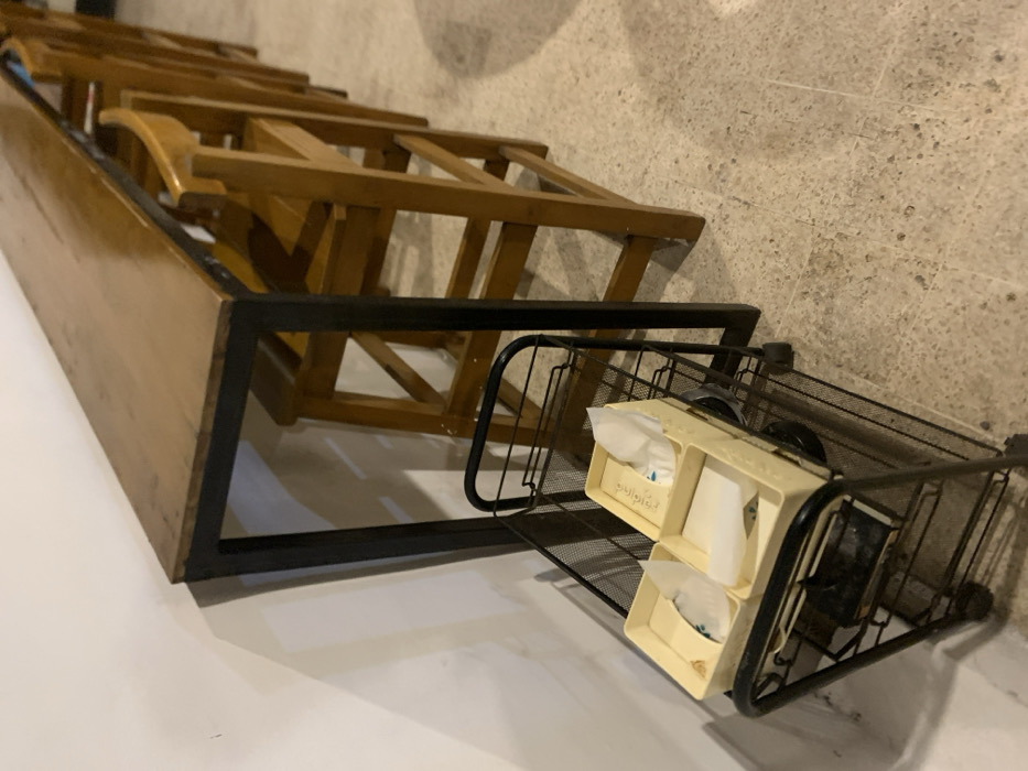 The image shows a wooden table and four wooden stools in a cafe in Probolinggo, Indonesia. The table and stools are made of light brown wood. The table has a black metal frame. The stools are stacked on top of each other. There is a black metal cart with a wire mesh shelf in the lower left corner of the image. The cart holds four small boxes filled with napkins and some books. The floor is tiled in beige tiles. The scene is empty and appears to be in the back of the cafe. The coordinates (-7.751, 113.213) correspond to a location in Probolinggo, Indonesia.