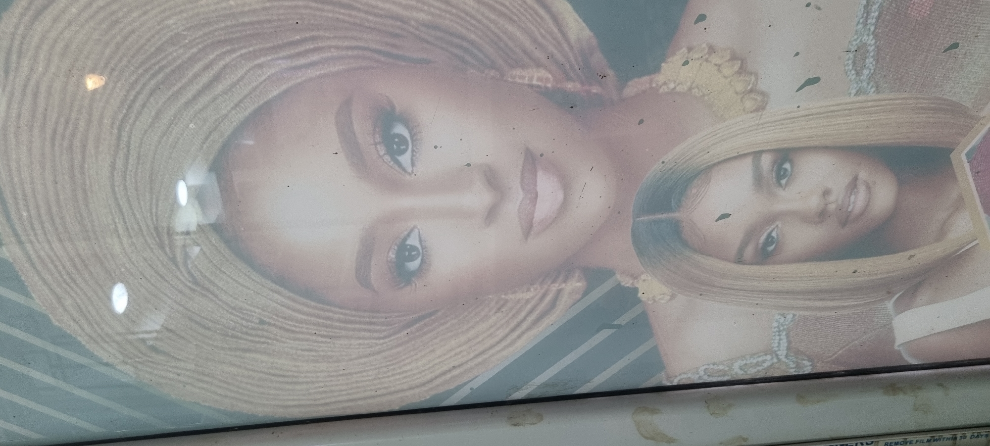 The image shows a poster with two portraits of a woman with light skin, likely an advertisement. The larger portrait features the woman wearing a wide-brimmed straw hat and yellow clothing, while the smaller portrait shows her with blonde hair and a white top. The poster appears to be affixed to a surface, potentially a building or vehicle, and is located in Lagos, Nigeria. This is inferred from the text "Remove film within 30 days" on the bottom right. The poster has some smudges and imperfections.