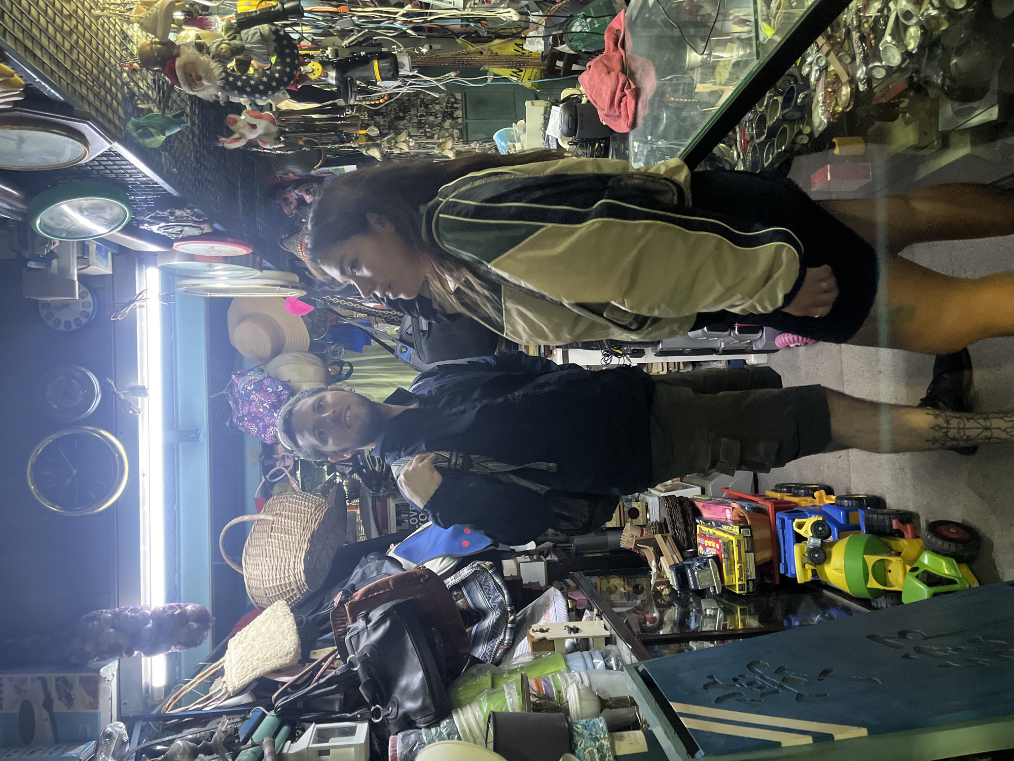 Two people are browsing inside a cluttered shop. A man with short blond hair is facing the camera and smiling. He is wearing a dark jacket and light green shorts. A woman with long dark hair is wearing a jacket with light and dark color blocking. She is looking into a glass display case filled with various items. The shop is packed with goods including bags, baskets, toys, clocks, and other miscellaneous items. The location is likely a vintage or antique shop in Buenos Aires, Argentina.