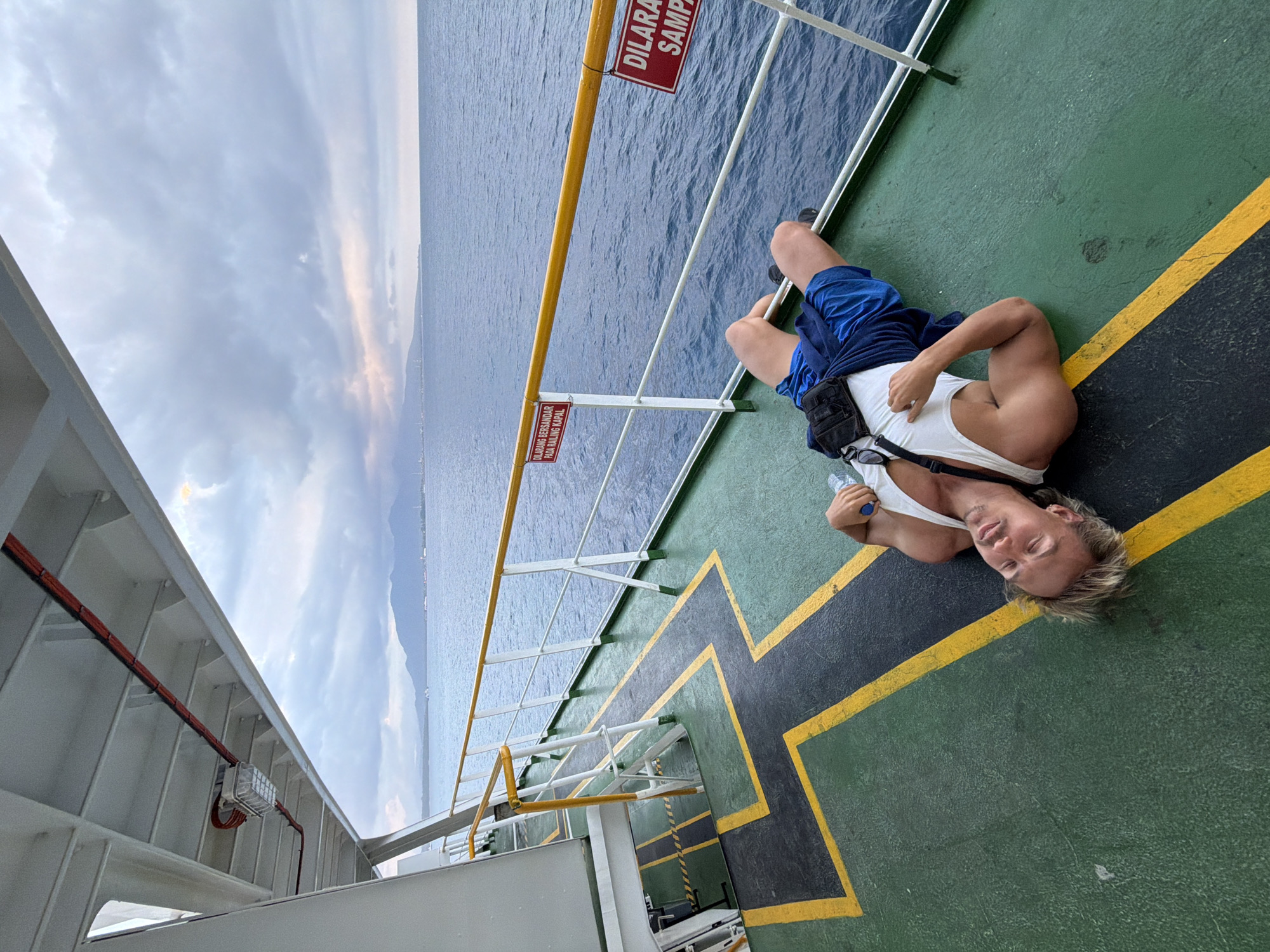A man lies on his back on the green deck of a ferry with yellow markings. He is wearing a white tank top and blue shorts. A black pouch is attached to his shorts. The man is holding a clear plastic water bottle in his right hand. The sea and a distant coastline with mountains are visible in the background under a cloudy sky. Two red signs with white text are affixed to the railing of the ferry. The sign on the left reads "DILARANG BERSANDAR PADA RAILING KAPAL", and the sign on the right reads "DILARANG SAMPA". The lighting suggests it is either early morning or late afternoon.