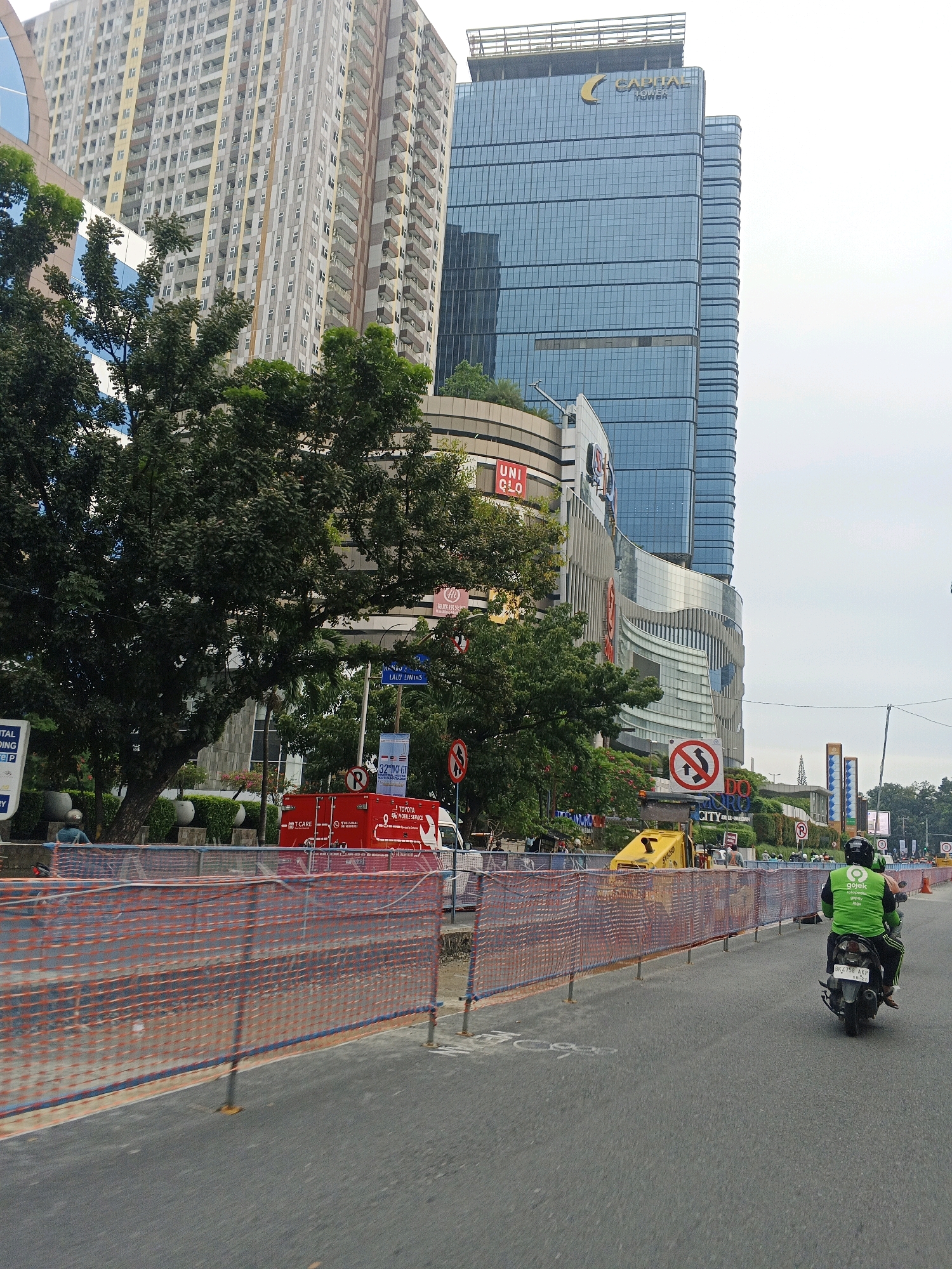 In City of Medan, Indonesia, a motorcycle rider wearing a green jacket with "gojek" branding is seen from behind, riding on a street. The rider is on a black motorcycle with a license plate visible. The street is lined with temporary orange safety fencing, suggesting ongoing construction or maintenance. In the background, several modern buildings dominate the cityscape, including a tall glass-fronted skyscraper with "CAPITAL TOWER" signage and another large building with a curved facade featuring a "UNIQLO" sign. Trees are present in the mid-ground, partially obscuring some of the buildings. Various traffic signs, including a "no overtaking" sign and a parking sign, are visible along the roadside. A red truck with "T-CARE" and "TOYOTA MOBILE SERVICE" is parked on the side of the road, and a yellow piece of construction equipment is also present. The sky is overcast.