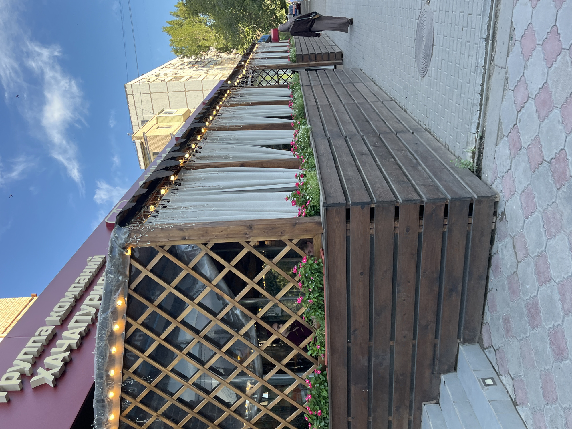 In Astrakhan, Russia, a street-level view captures the exterior of a restaurant with an outdoor seating area. The building facade, painted a deep red, features raised white lettering that reads "ГРУППА КОМПАНИЙ ХАЧАПУРИ" (Group of Companies Khachapuri). An awning extends from the building, covered with clear plastic and adorned with string lights and festive white curtains. Beneath the awning, a wooden lattice structure encloses the seating area, which is decorated with rows of blooming pink flowers. A person wearing a brown hijab and long dress walks away from the camera on a patterned brick sidewalk. The environment is urban, with apartment buildings and trees visible in the background under a partly cloudy blue sky.
