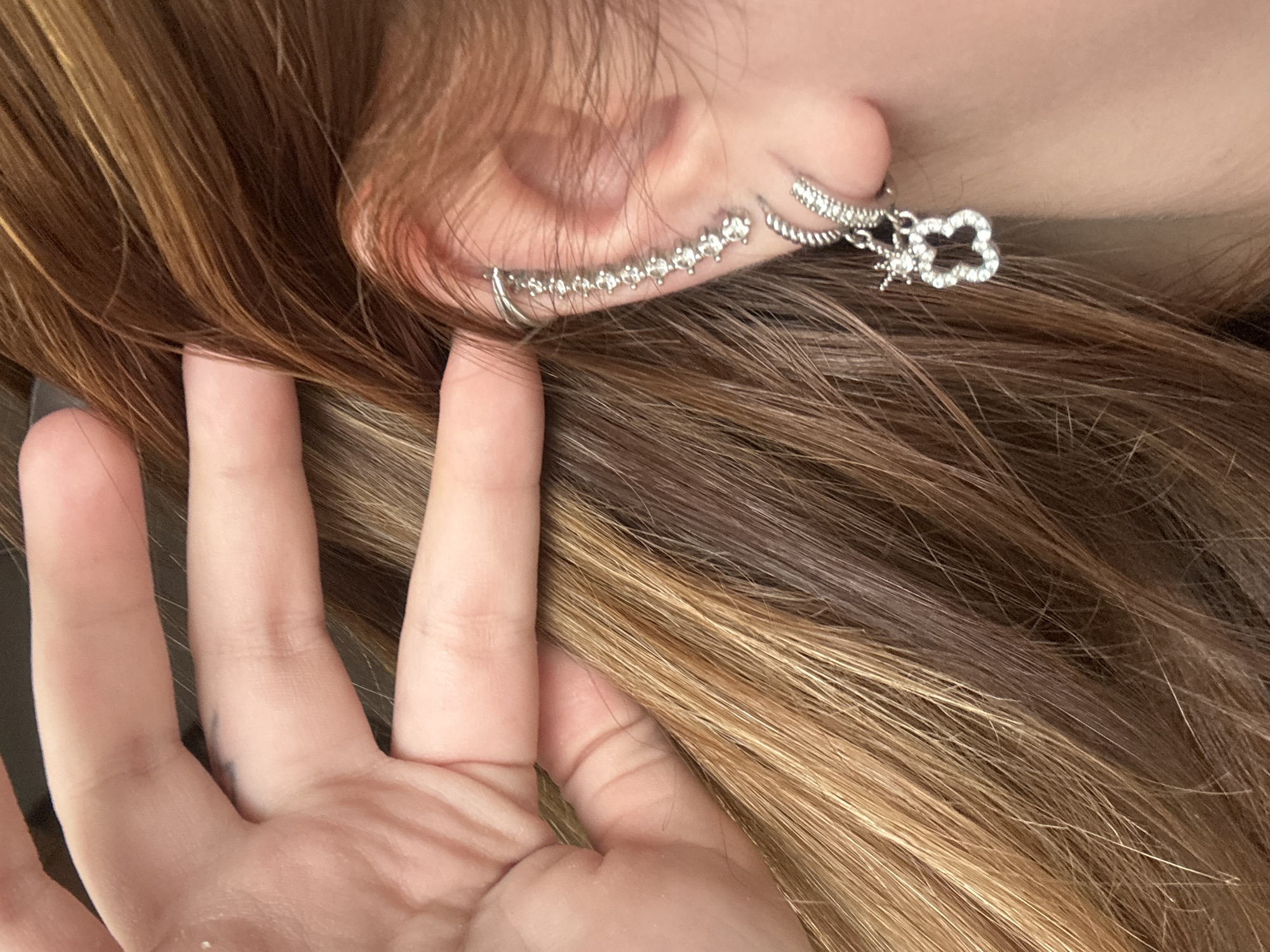 This close-up shot captures a partial view of a person, focusing on their right ear, strands of hair, and a hand. The person has long, wavy hair, primarily dark brown with prominent lighter blonde or caramel highlights, creating a streaked effect.
Adorning the ear are multiple silver-toned earrings. A distinctive ear cuff or climber, studded with small, clear rhinestones, follows the curve of the outer ear cartilage. Below it, a small, simple silver hoop earring is visible in the lobe. Further back, a dangle earring hangs, featuring a four-leaf clover or flower-shaped charm also encrusted with clear stones, and a smaller star-like charm attached.
A light-skinned hand, likely the person's left hand, is positioned in the foreground, with its fingers gently pulling back or holding strands of hair, revealing the ear and its adornments. The scene appears to be an intentional pose to showcase the hair and earrings.
The lighting suggests an indoor setting or a brightly lit outdoor area, but the specific time of day is indeterminate. There are no explicit background details to indicate a particular location within Troyan, Bulgaria, beyond the provided context. No visible text is present in the image, and emotions are not discernible from the visible portions of the subject.
