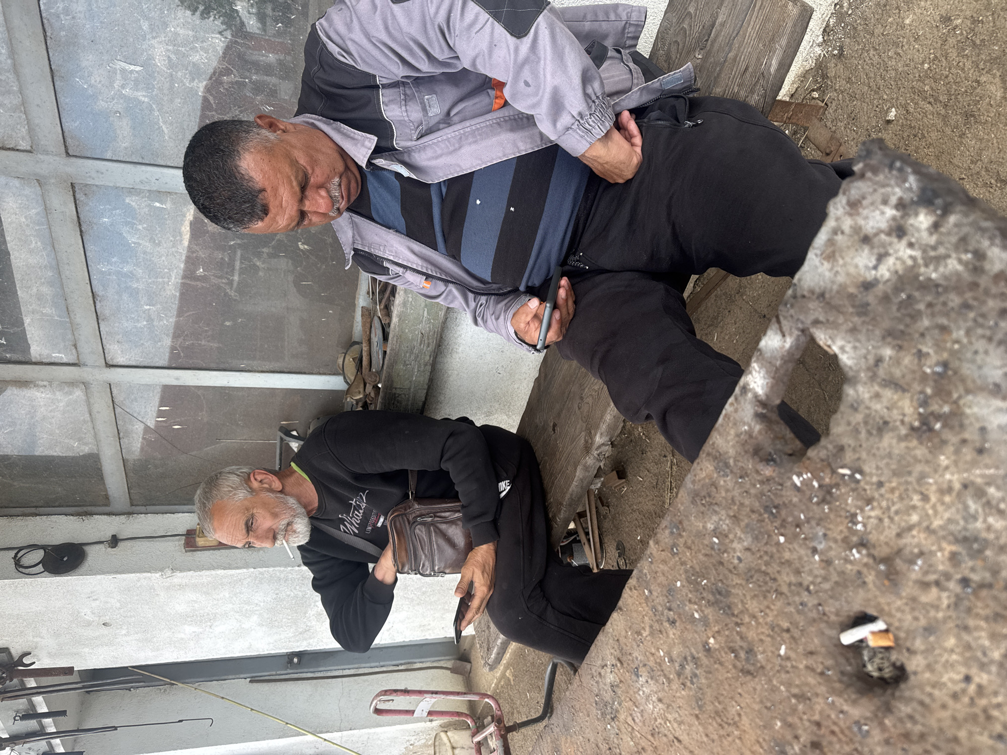 The image depicts two men seated in an outdoor setting, possibly a workshop or industrial area, in Sarnevo, Bulgaria. One man is wearing a gray and black jacket with a striped shirt and black pants. He is seated and looking down, holding a dark-colored rectangular object in his hands. The other man is wearing a black sweatshirt, black pants with a visible Nike logo, and a brown leather shoulder bag. He is smoking a cigarette. In the foreground, there is a rough, weathered surface with cigarette butts scattered on it. Various tools and equipment can be seen in the background, along with a concrete structure and a partial view of a metallic roof.