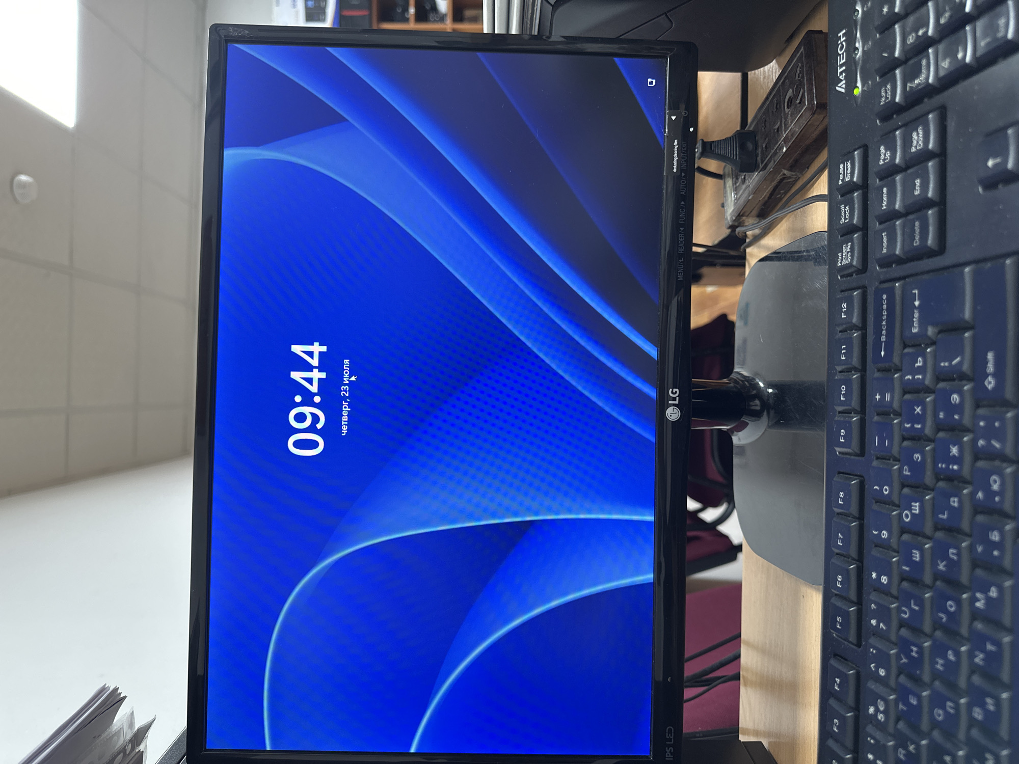The image shows a computer monitor displaying a lock screen with the time "09:44" and the date "четверг, 23 июля" (Thursday, July 23) in Russian. The monitor is an LG IPS LED model. Below the monitor is a black A4TECH keyboard. The scene appears to be an office or study environment, with a wooden desk and some office supplies visible in the background, including stacks of paper and a power strip. The lighting suggests an indoor setting. There are no people visible in the image. The image does not provide any specific information about the location being Kadamjay, Kyrgyzstan.