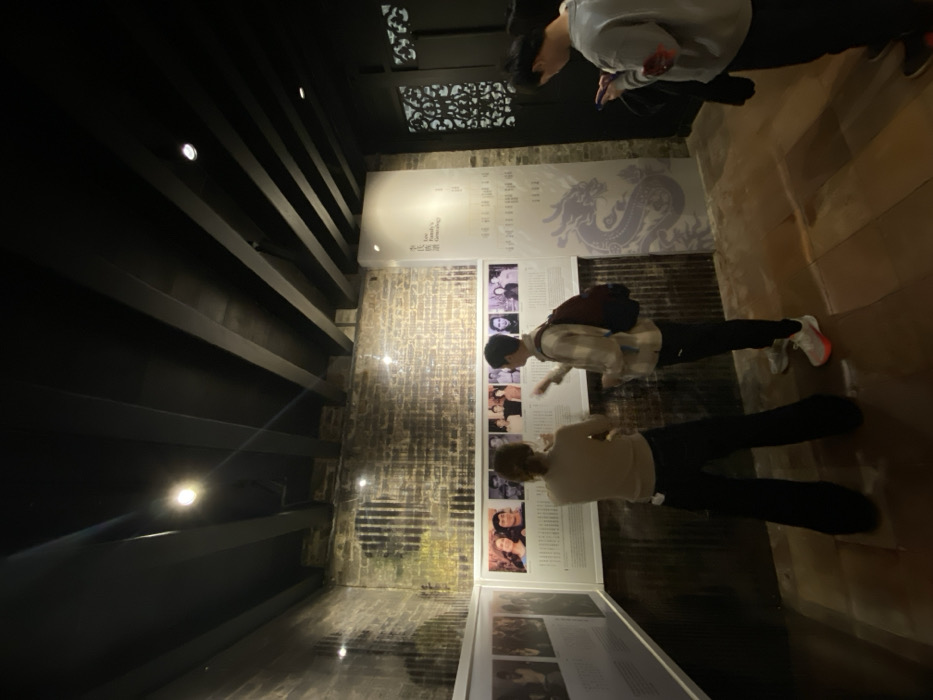 This media file depicts an indoor scene within what appears to be a museum or exhibition space, likely located in Guang Zhou Shi, China. Three individuals are present, observing various display panels.
The setting features a ceiling composed of dark, exposed wooden beams with integrated spotlights, and decorative patterned panels are visible towards the upper back of the room. The floor is made of polished wood, reflecting some of the overhead lighting.
Along the right side of the image, several display panels are arranged. A large white panel at the back features a subtle light purple dragon motif, Chinese characters, and partial English text that appears to say "On Family V... Genealogy." Below this, a series of smaller white panels present rows of black and white photographs of individuals, each accompanied by descriptive text.
In the mid-ground, a person wearing a plaid shirt and dark pants is leaning forward, engrossed in an exhibit. In the foreground, another individual wearing a light-colored top and dark pants is also focused on the displays. Further back, a third person in a light grey hoodie stands, seemingly interacting with a mobile phone.
The left wall of the room has a textured surface, possibly brick or distressed plaster, which is illuminated by spotlights. The overall lighting is artificial and somewhat subdued, creating a focused atmosphere for viewing the exhibits. The people appear calm and engaged in their activity.