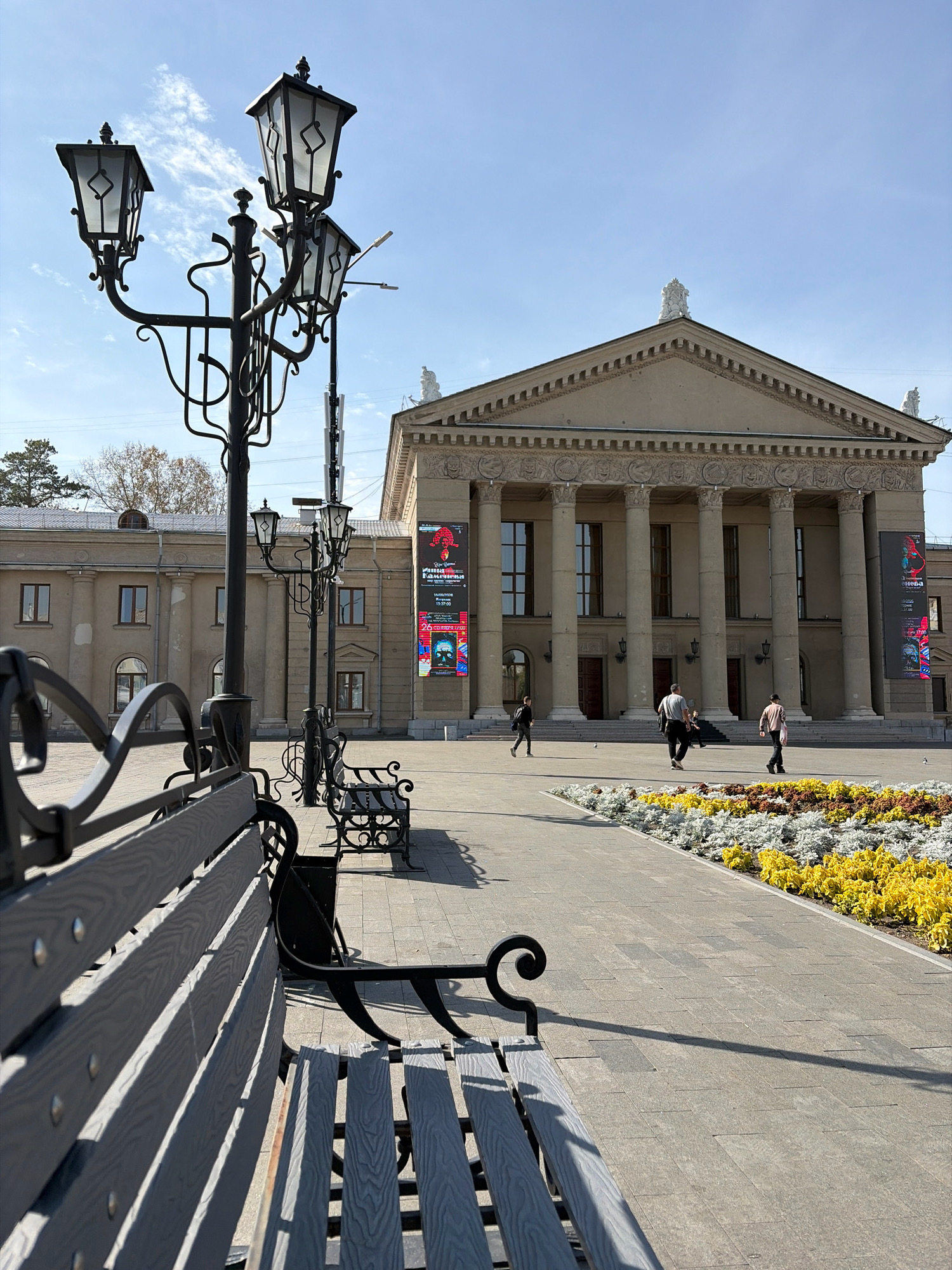 The image captures a daytime scene in front of a grand building with a portico of large columns, suggesting a public institution or theater, possibly located in Angarsk, Russia. The building's facade is light-colored and features decorative elements above the columns. Two large banners hang vertically on the building's facade, displaying posters with text and graphics.
In the foreground, a black wrought-iron bench with gray wooden slats is visible, casting a shadow on the paved ground. Further back, another similar bench is present. A tall, ornate lamppost with three lights stands to the left of the frame, also casting a shadow. Another lamppost is visible behind it.
A paved walkway runs alongside a vibrant flower bed filled with yellow and white flowers. Several people are walking in the middle ground: one person in a dark jacket and pants is walking towards the left, another person in a gray shirt and dark pants is walking towards the right, and a third person in a light-colored shirt and dark pants is also walking towards the right. The sky is clear blue with a few wispy clouds. The environment appears to be an urban plaza or square.