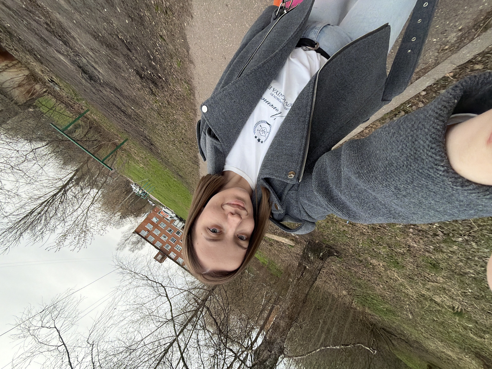 An image, rotated ninety degrees to the left, captures a young woman outdoors in what appears to be Reshetnikovo, Russia. The woman, with medium-length brown hair and light eyes, is centered in the frame, looking directly at the camera with a neutral to slightly pleasant expression. She is wearing a grey zip-up jacket over a white t-shirt, and light-colored pants, likely jeans, secured with a black belt.
On her white t-shirt, visible text reads "ORIGINAL DESIGN BY" and "DVIGUYA E" (partially obscured), along with a word that appears to be "Movenne" or "Mavenne." Below this text is a logo resembling a crest or shield featuring stars and a crown-like symbol.
The background suggests a late winter or early spring setting. Many trees are bare of leaves, though hints of green grass are visible on the ground. A dirt or gravel path runs alongside the woman. In the distance, a multi-story brick building with numerous windows is visible, typical of residential architecture. A green metal structure, possibly part of a fence or playground equipment, is also in the background. The sky is overcast, indicating a cloudy day. The overall scene suggests a casual outdoor moment in a semi-urban or suburban environment.