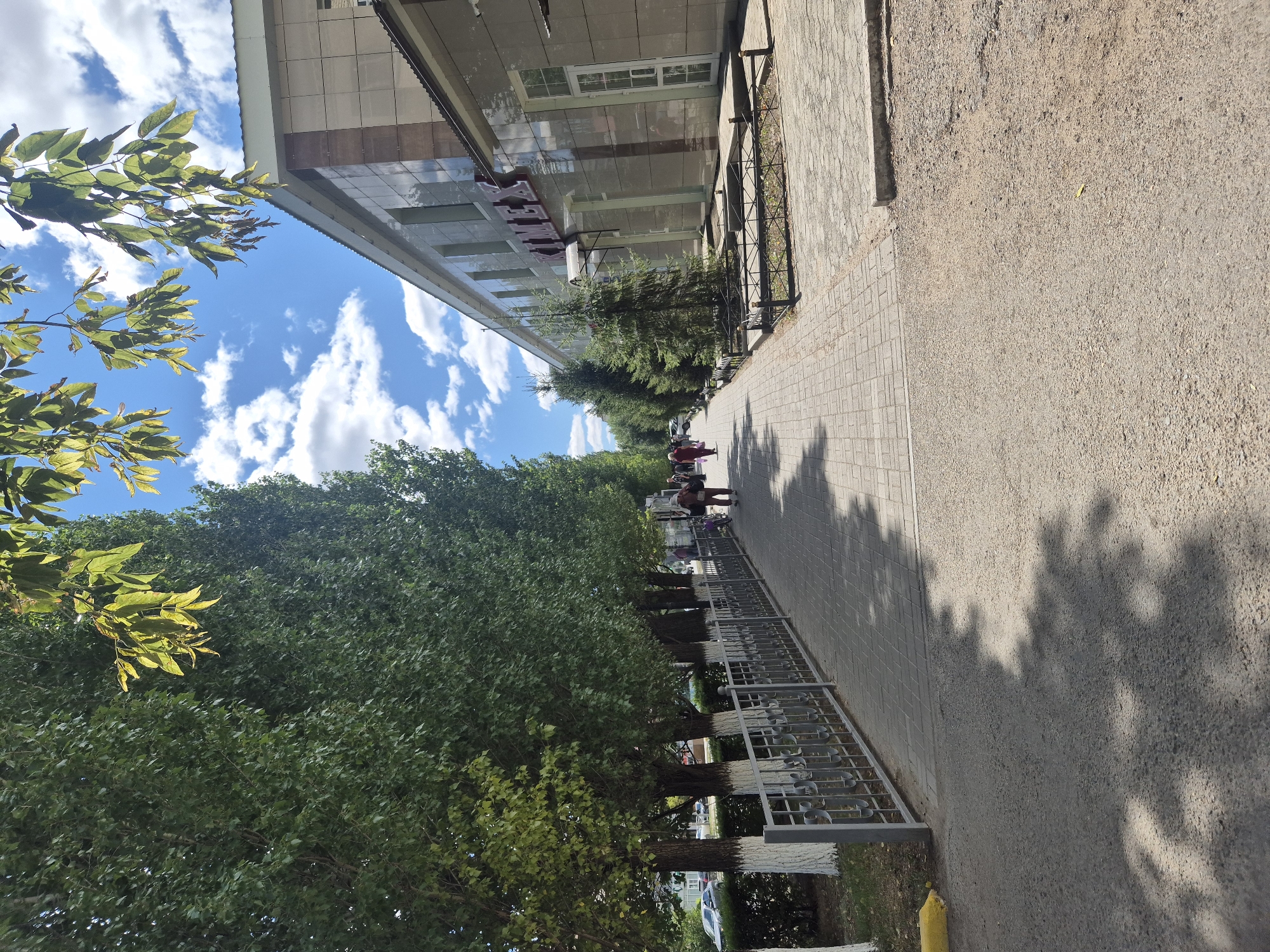 The image shows a paved pedestrian walkway alongside a row of tall, leafy trees with their trunks painted white. A metal railing separates the walkway from a grassy area with more trees and bushes. On the right side of the walkway is a modern building with a tiled facade. The building has large windows and a sign with red lettering that appears to read "KIMEK." Several people are walking along the sidewalk in the distance. The sky is blue with scattered white clouds. The scene depicts a street in the city of Kokshetau, Kazakhstan.