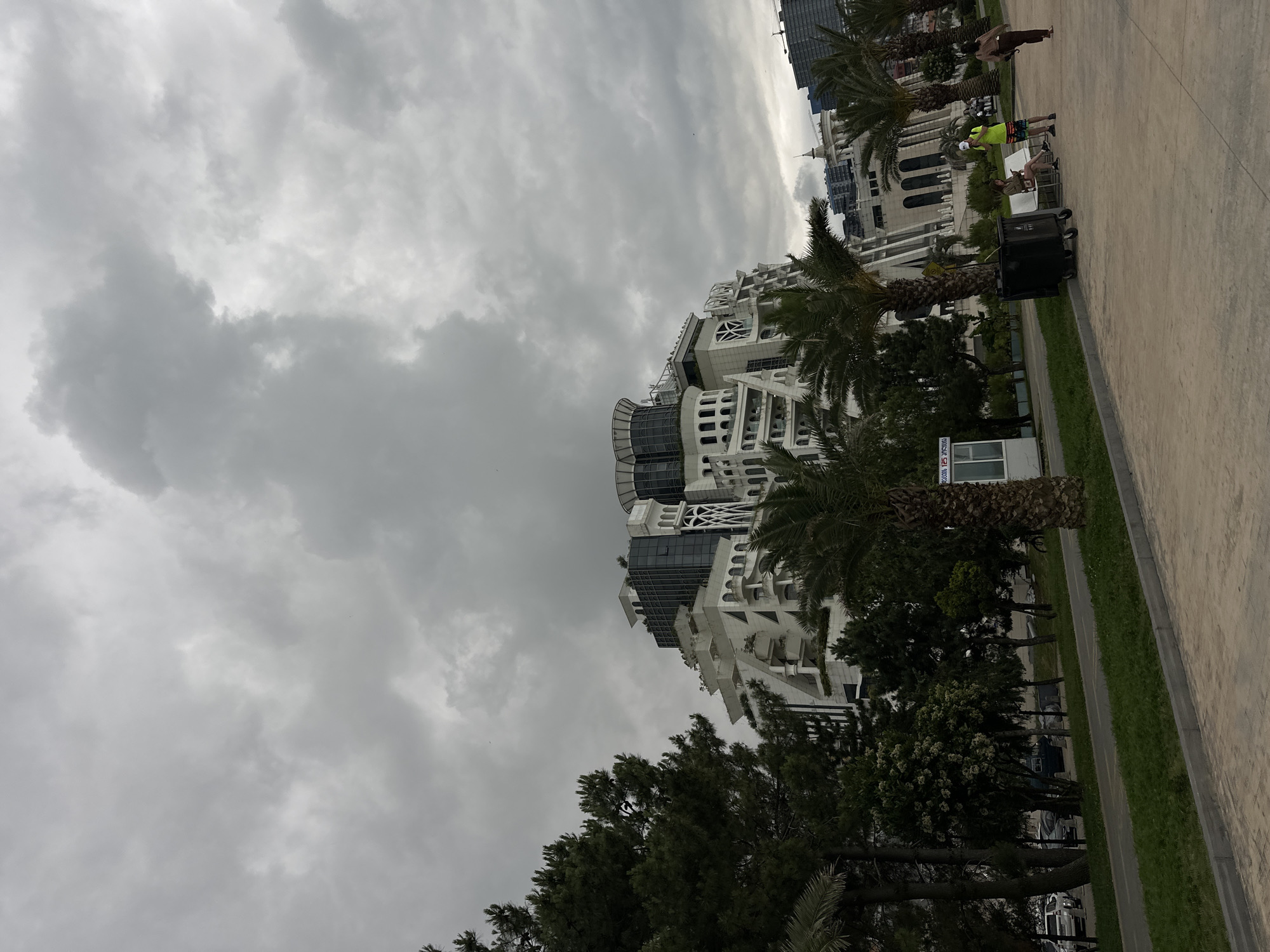 The image depicts a cloudy day in Batumi, Georgia, with a focus on the striking architecture of the Buildings. The main structure is a white, multi-tiered building with numerous balconies, intricate white decorative elements, and glass-paneled upper floors. To its left, a dense cluster of dark green trees, including pine trees, partially obscures the view of the building. Palm trees are scattered throughout the foreground and mid-ground.
In the foreground, a paved walkway runs parallel to a grassy area and a road. Several vehicles are visible parked along the roadside, including a white SUV. A small white kiosk stands near the palm trees.
Further into the scene, a black trash bin with wheels is positioned on the pavement. Two people are seated on a white bench near the trash bin: one person wearing a lime green shirt and patterned shorts is standing and looking at their phone, while another person sits on the bench looking at their phone. A third person, wearing a brown top and pants, walks away from the bench towards the right side of the image. In the far background, other buildings and a construction crane are visible. The sky is heavily overcast with dark gray clouds. The overall scene suggests an urban environment with a mix of natural elements and modern architecture.