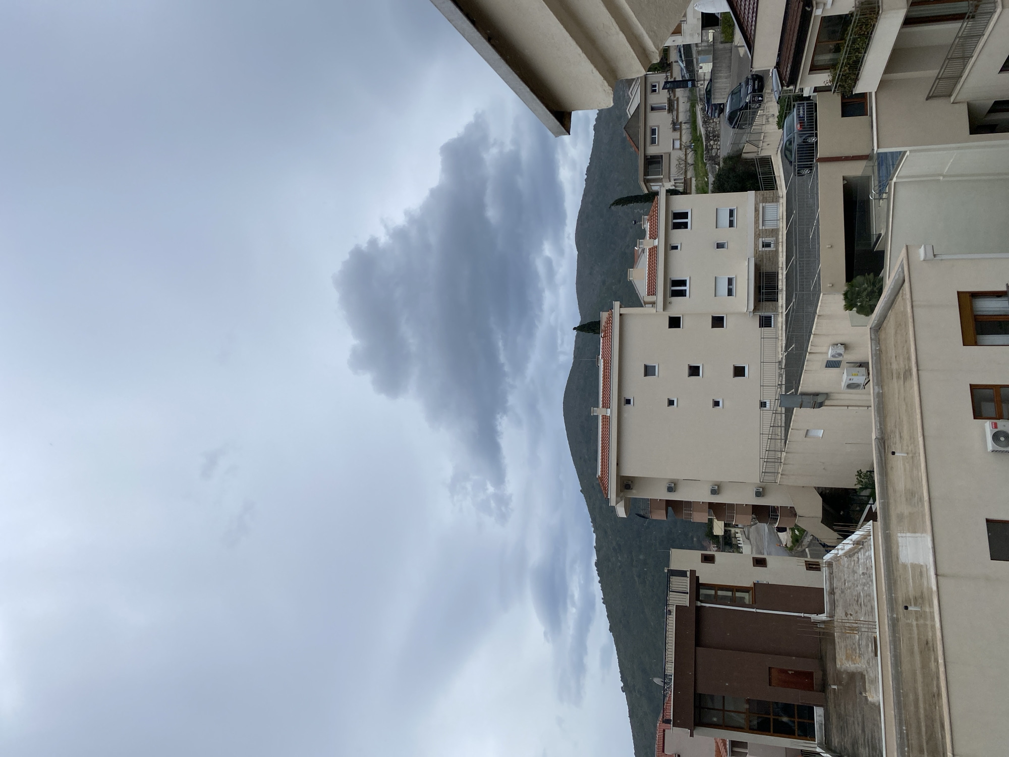 The image shows an overcast sky with a large gray cloud formation above a hillside in Budva, Montenegro. Buildings of varying heights and styles are clustered on the hillside, with a road visible between them. Cars are parked along the road. The photograph appears to have been taken from an elevated position, possibly a balcony or rooftop. The foreground features architectural elements, indicating the vantage point is within a building. Air conditioning units are affixed to the exterior of several buildings.