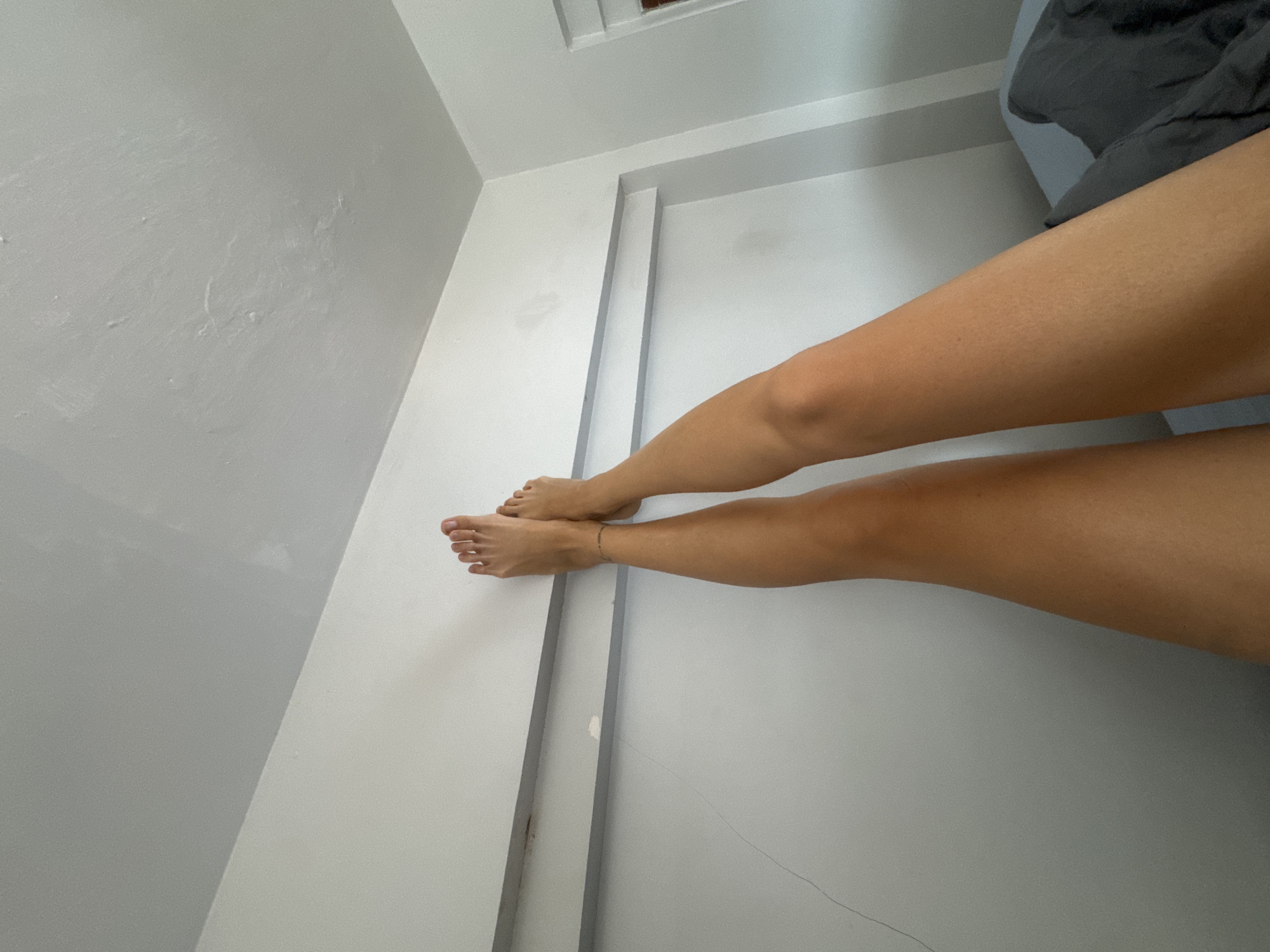 The image shows the lower body of a person, focusing on their legs and feet. The person has tan skin and appears to be barefoot, wearing a thin anklet. Their legs are positioned horizontally across the frame, with the feet closer to the viewer. They are resting against a white, textured wall, which has some subtle imperfections and stains. To the right of the legs, a dark gray fabric, likely bedding, is visible, suggesting the scene is indoors, possibly in a bedroom. The lighting is soft, indicating it could be daytime, but there are no clear indicators of the time of day or weather. The overall mood is relaxed and casual. The setting appears to be a room with white walls and a lighter gray wall in the background, with some architectural details like recessed trim. There is no visible text in the image. The location context is North Kuta, Indonesia.