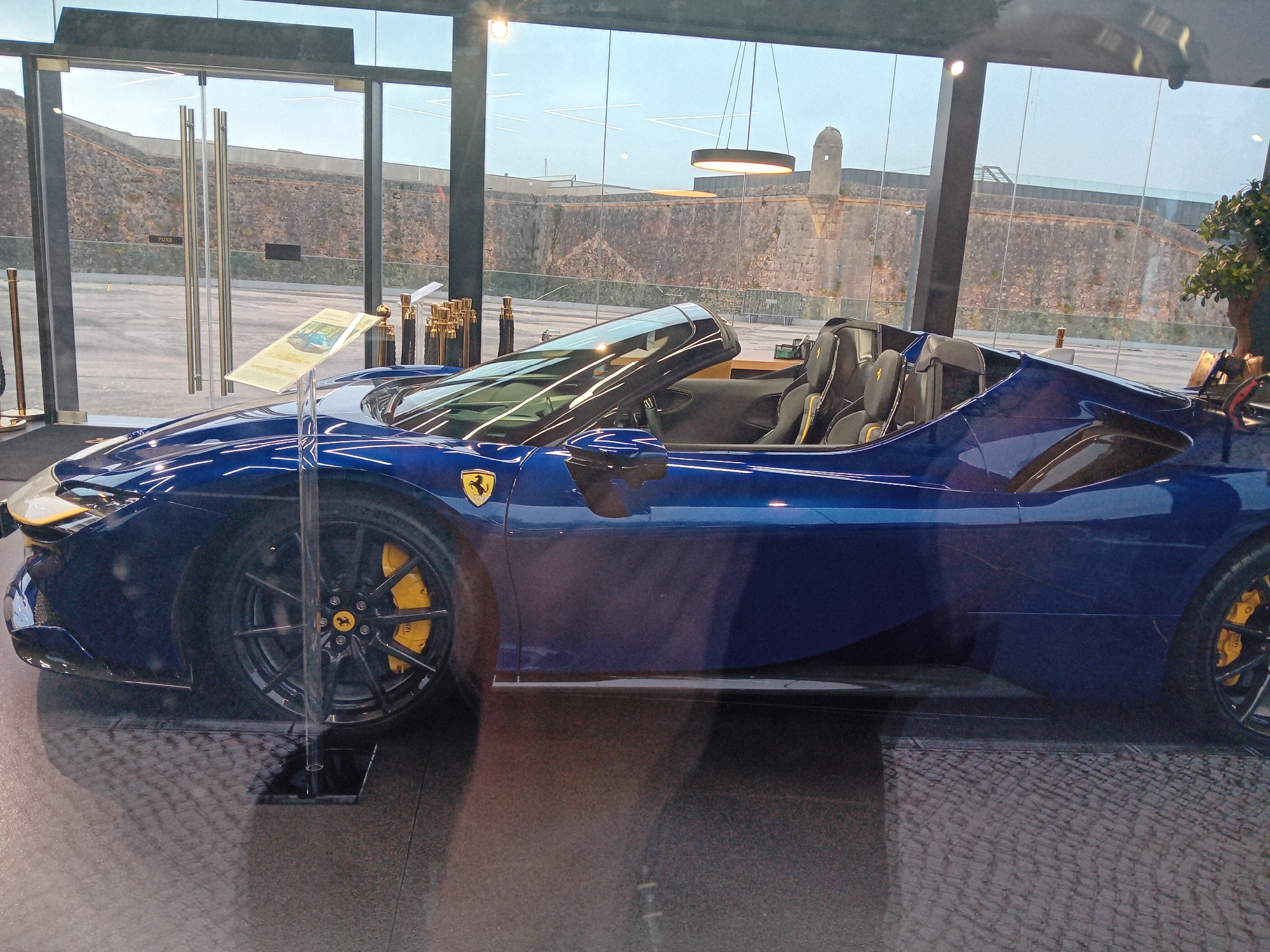 The image shows a blue Ferrari Monza SP1/SP2 on display inside a modern building with large glass windows. The exterior of a fortress-like structure is visible through the windows. This scene is likely in Cascais, Portugal, due to the presence of the fortress, which resembles the Citadel of Cascais. The car sits on a dark floor. A display stand with printed material is situated next to the car, and ornate gold-colored poles are lined up beside it. No people are visible in the direct frame, though reflections on the glass suggest a person taking the picture. The overall lighting is bright and airy.
