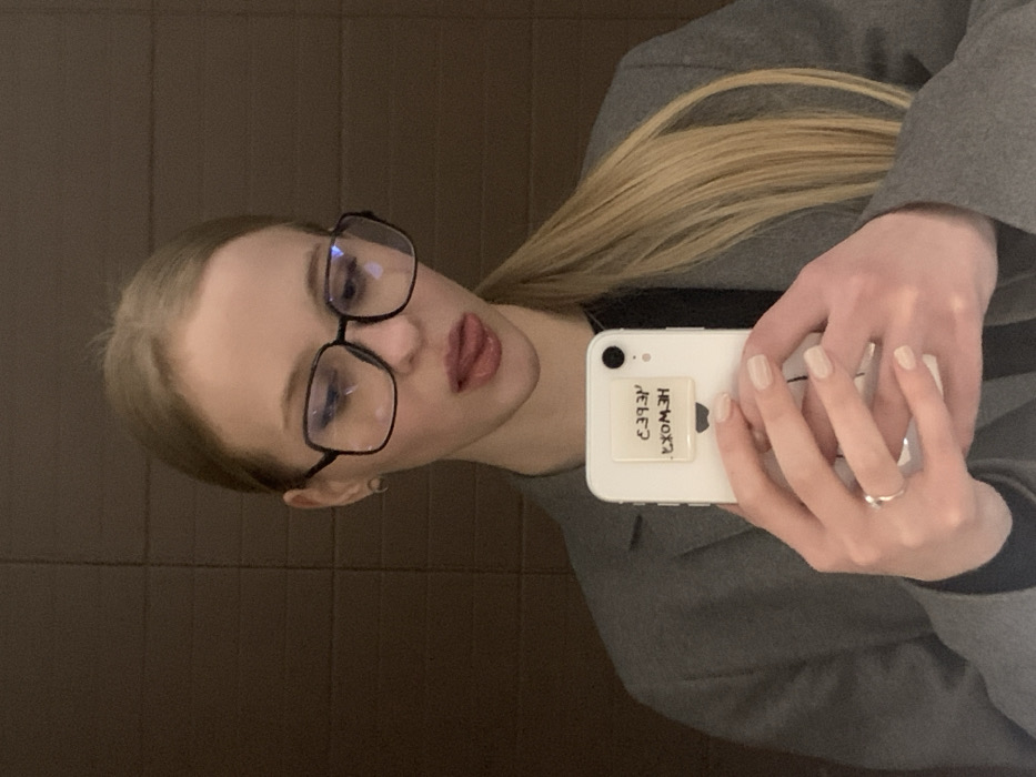 A selfie of a woman with blonde hair pulled back in a ponytail. She wears large, dark-framed glasses. She pouts slightly, sticking her tongue out. She holds a white smartphone with a sticker that reads "He works?" but it's mirrored and upside down. She wears a gray jacket or blazer. The background is a dark brown, possibly a wall of some sort.