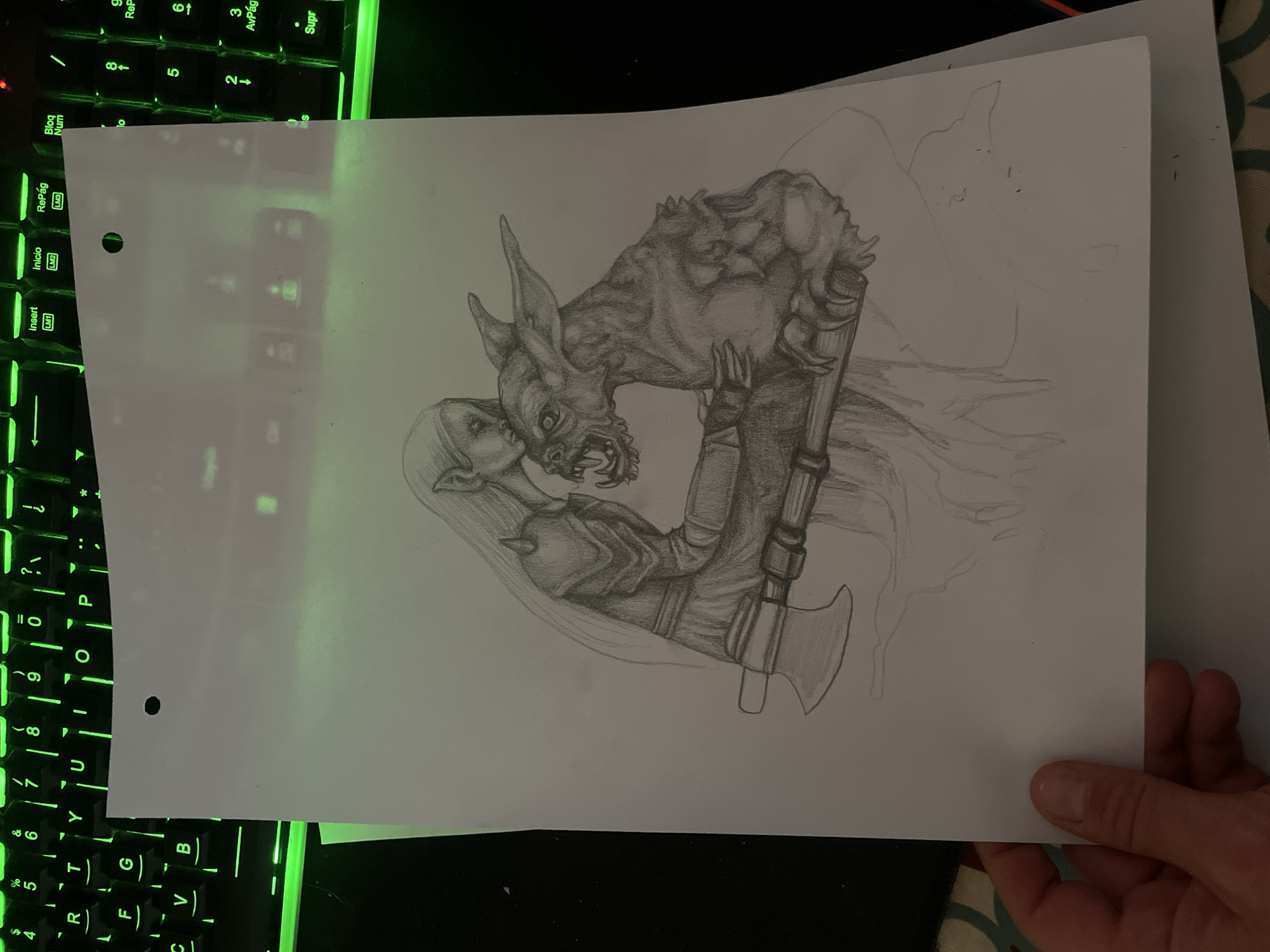 A pencil drawing on a sheet of paper is presented in the image. The drawing depicts a fantasy scene with two primary figures: a female elf-like character and a monstrous creature. The female character appears to be embracing or kissing the creature, which has a snarling expression and pointed ears. The creature is wearing some kind of armor or clothing and wields an axe. The background of the drawing is only partially rendered, with light pencil strokes suggesting an undefined environment. The drawing style is detailed, with shading and cross-hatching to create depth and texture. The sheet of paper with the drawing is situated on top of a backlit keyboard with green lighting. The hand of a person is visible holding the paper.