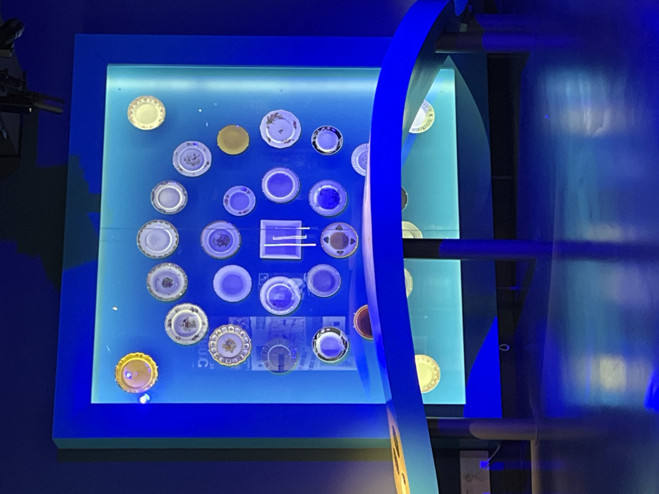 The scene depicts an illuminated exhibition display located within the Cité Internationale de la Gastronomie et du Vin (CIGV) in Dijon, France. A large, rectangular display case dominates the center, framed in deep blue. Its interior features a back panel emitting a gradient of cyan to light blue illumination. Mounted on this panel are approximately 25 to 30 diverse plates, predominantly circular, varying in size, color, and decorative patterns. Most plates appear blue or bluish-white due to the intense blue lighting, though several retain warmer tones, notably a gold-colored plate at the top-left and another at the top-right. Faint text, including the numeral "500," is partially visible behind some plates. In the foreground, a dark blue, curved, and undulating counter is supported by multiple dark vertical posts. The floor is dark and reflective, mirroring the blue light from the display. The immediate environment is dimly lit, with the display providing the primary light source. No individuals are present in the image.