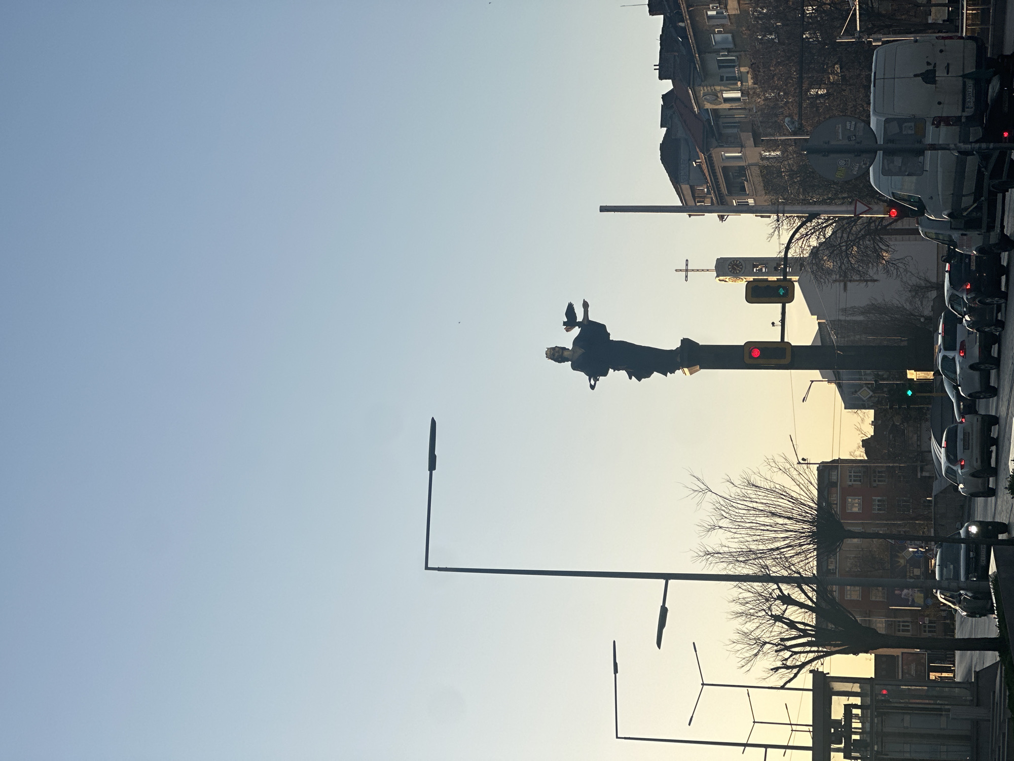 The image depicts the Statue of Sveta Sofia (Saint Sofia) in Sofia, Bulgaria. The statue stands atop a tall column, a bird perched on one of her hands. To the right of the statue is a street with cars and vans stopped at traffic lights. Buildings line the street, including some with visible architectural details and a clock tower with a cross atop it. Streetlights are visible in the foreground. The sky is light blue.