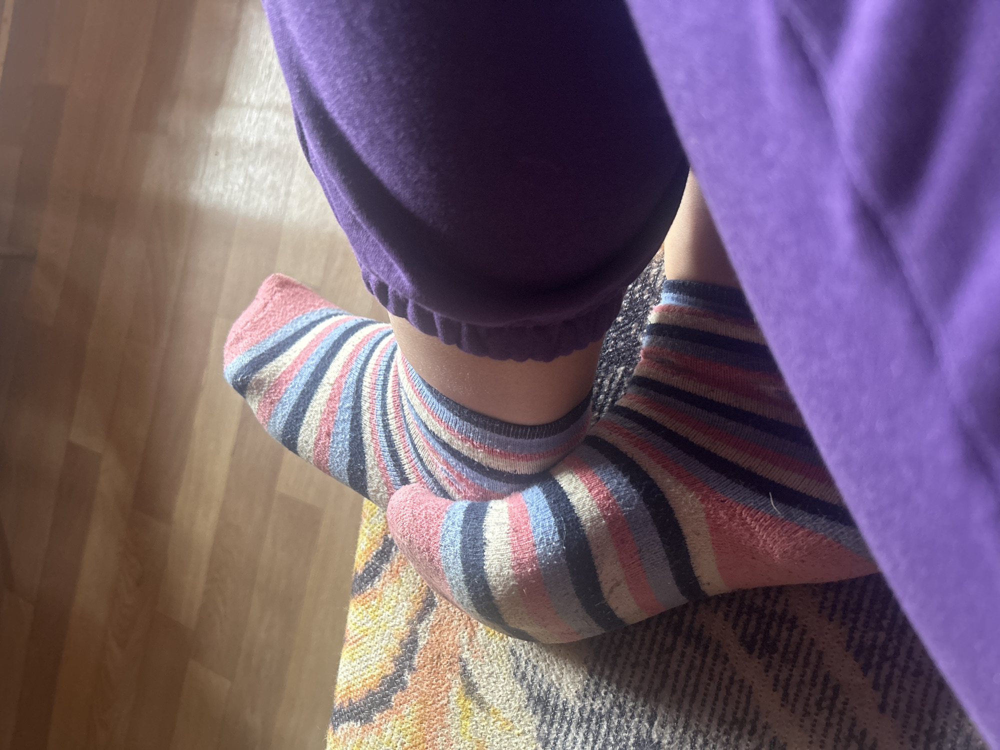 The image shows a close-up view of a person's feet, wearing striped socks and purple pajama bottoms. The socks have a pattern of pink, blue, white, and black stripes. The person's feet are resting on a textured surface, possibly a couch or blanket, which has a geometric pattern in shades of yellow, orange, and gray. In the background, a wooden floor is visible, with warm lighting suggesting an indoor setting. The scene appears to be casual and relaxed, perhaps during the day given the lighting. There is no visible text in the image.