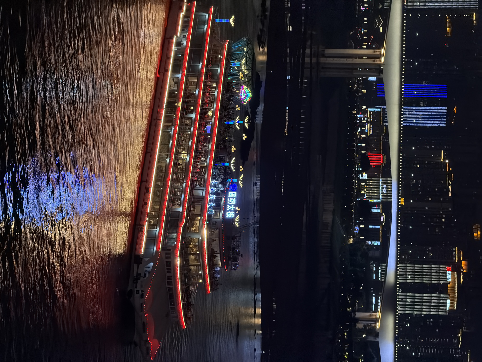 This is an overhead nighttime shot of a wide river with a large, multi-deck tour boat on the left. The boat is illuminated with red and white lights, highlighting its various levels and the people visible on board. The water reflects the boat's lights and the city lights from the shore.
On the right side of the image, a city skyline is visible, with numerous buildings lit up in various colors, including blue, white, and red. A distinctive white, curved structure, possibly a bridge or a building feature, dominates the center-right of the frame. The overall scene suggests a vibrant urban environment at night, with the river acting as a central element.
Text is visible on the boat, appearing to be in Chinese characters: "国药大药" and what looks like a logo with a sun-like element. The city lights suggest the time is night, and the clear reflections in the water indicate calm weather. The atmosphere is lively, with the illuminated boat and city suggesting a busy tourist or entertainment area.