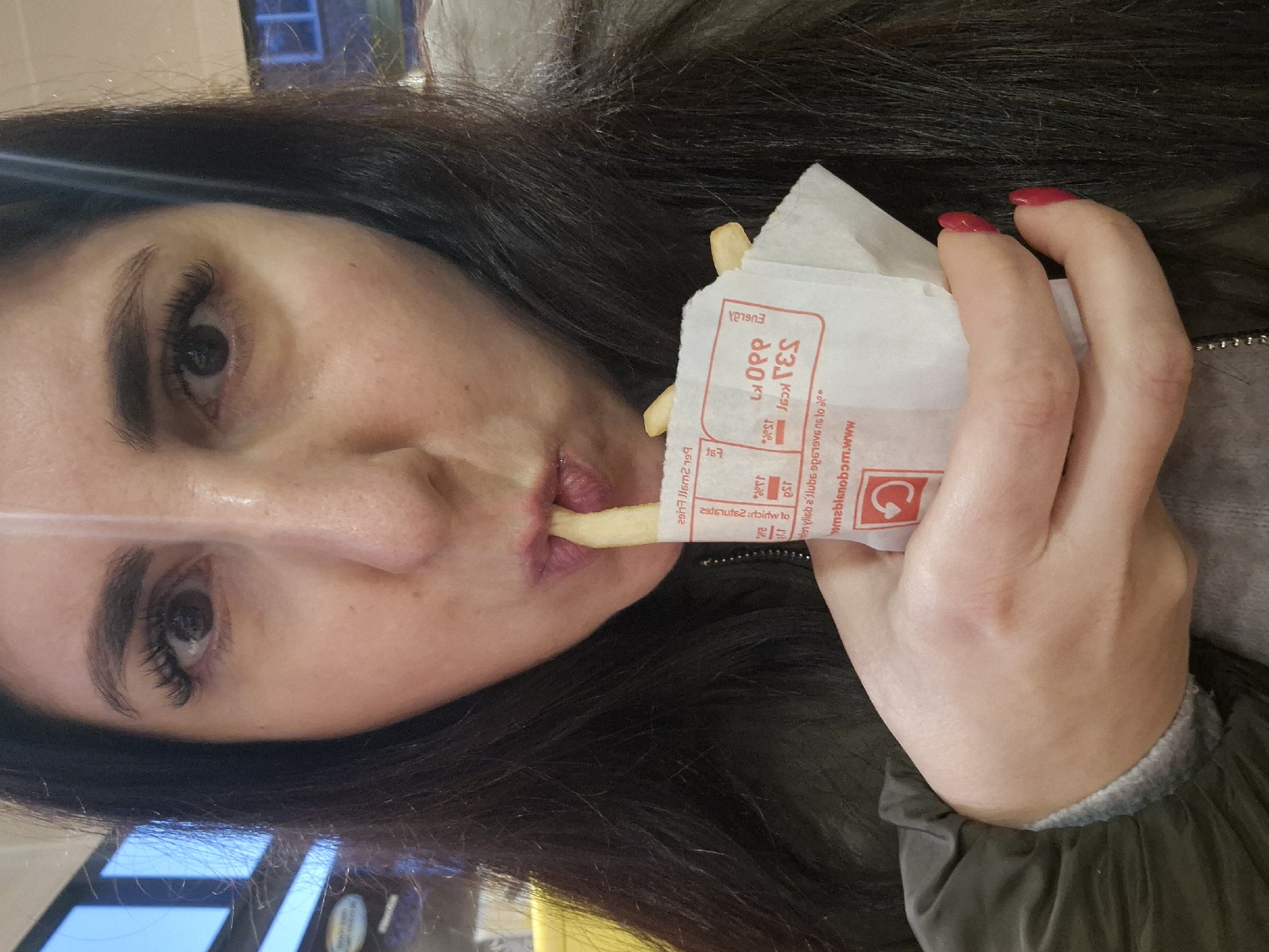 The image depicts a woman with long, dark hair and red nail polish on her fingers. She is holding a packet of French fries, and she is in the process of eating one. The text on the packet appears to be partially reversed, but includes text like "bloom.www," calorie information "330 kcal," and the word "EUGLA." A portion of her clothing, possibly a jacket, can be seen, revealing a grey cuff and a dark green fabric. The scene appears to be indoors. The location is not discernable based on the provided information.