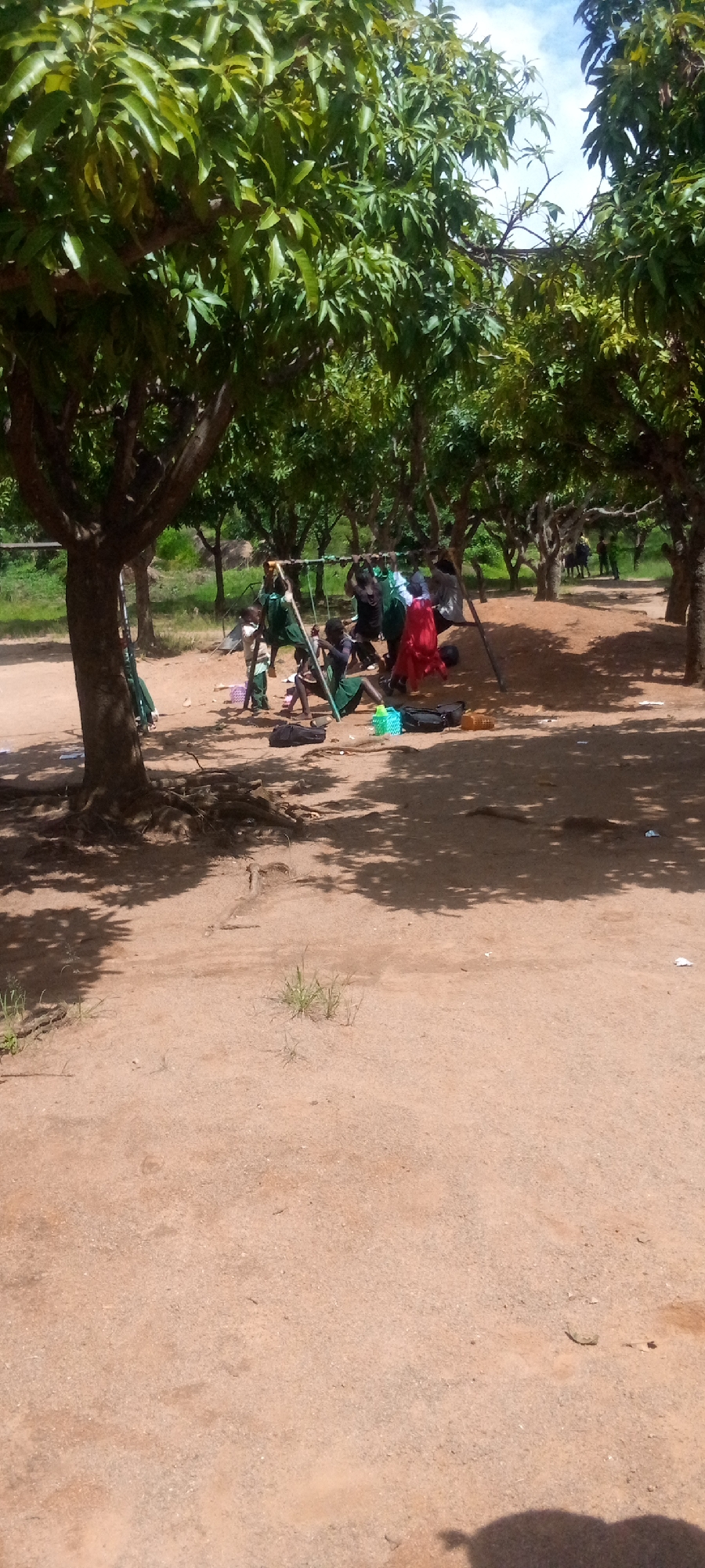 In this outdoor scene, a group of people, appearing to be children, are gathered under the shade of trees. They are near a rudimentary swing set, made of metal poles and a frame. Some of the children are seated on the ground, while others are standing. One child, wearing a red garment, is seated on the swing. Several bags and a few colorful plastic containers are scattered on the sandy ground. The trees have full green foliage, suggesting a warm climate, and the dappled sunlight indicates it is daytime, likely with clear skies. The ground is predominantly sandy with some sparse vegetation. The overall impression is of a relaxed gathering, possibly during a break or playtime. There is no visible text in the image.