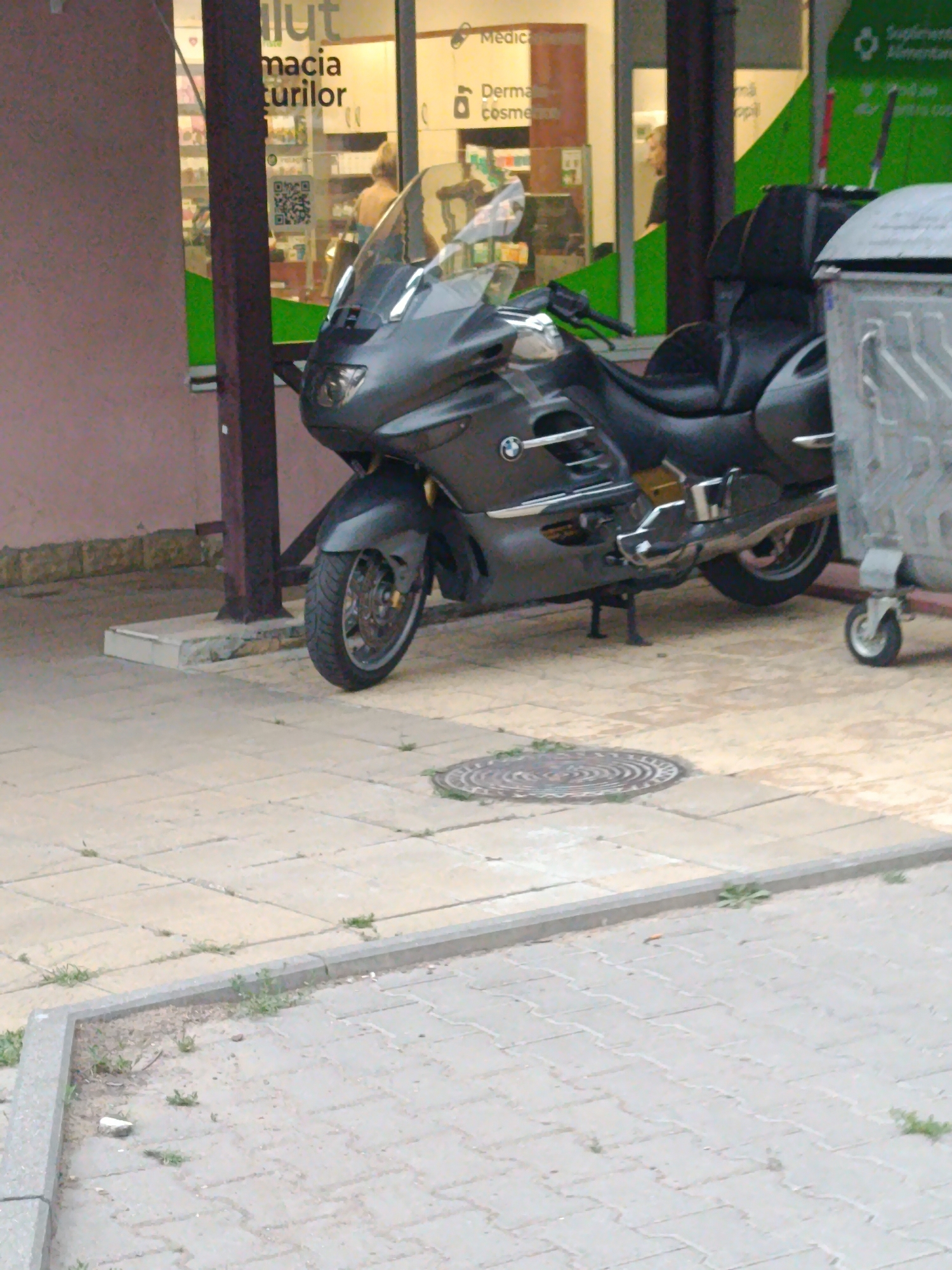 The image shows a grey BMW motorcycle parked on a paved outdoor area in front of a storefront. The storefront appears to be a pharmacy, with signage that includes "Farmacia Turilor" and "Medica". A large, silver trash receptacle with a wheel is visible to the right of the motorcycle. The ground is paved with square and rectangular tiles, and there are some weeds growing in cracks and along the edge of the pavement. In the background, through the storefront windows, shelves of products and people can be seen. The lighting suggests it is daytime.